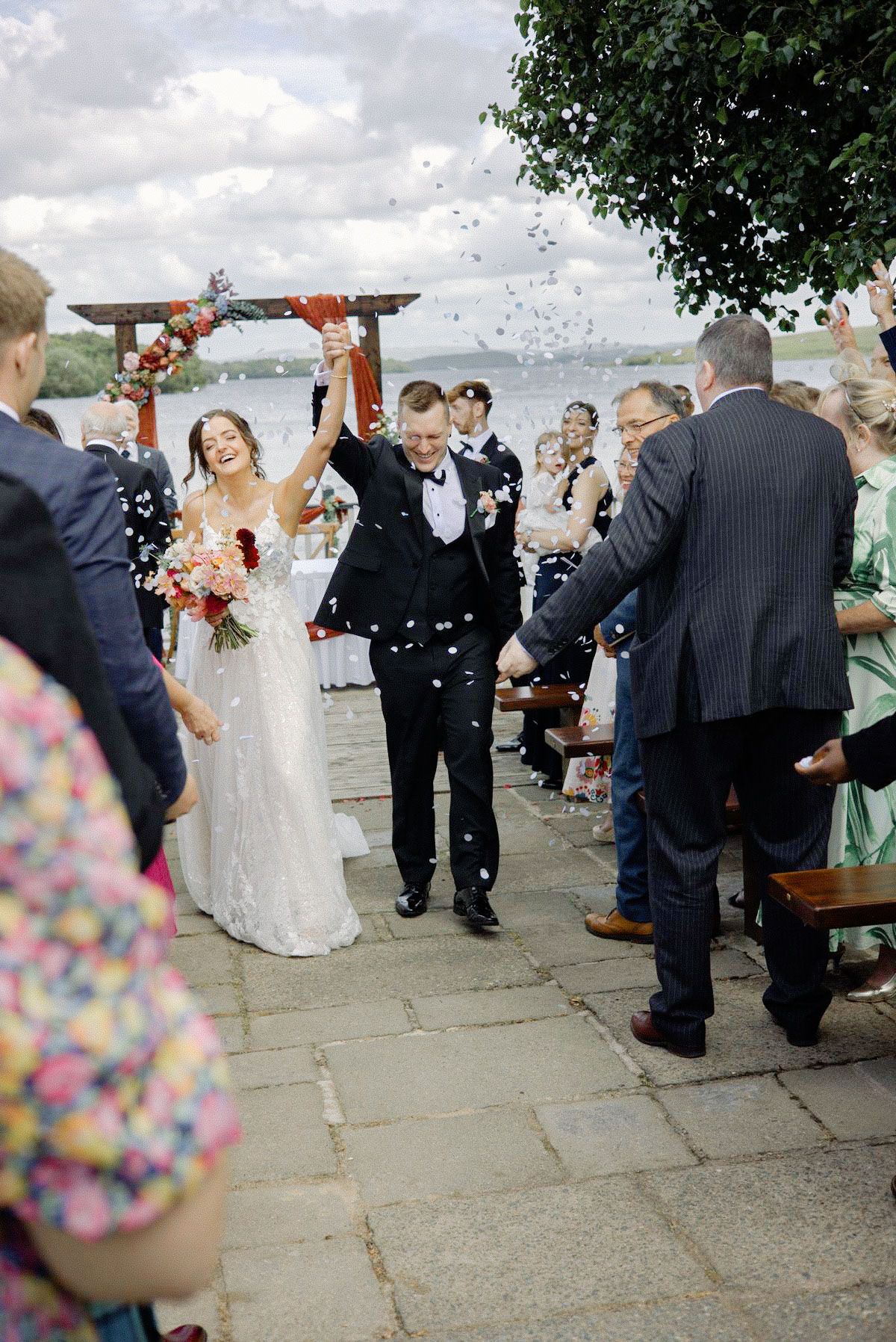 Joyful bride and groom walking through confetti shower after outdoor lakeside wedding ceremony.
