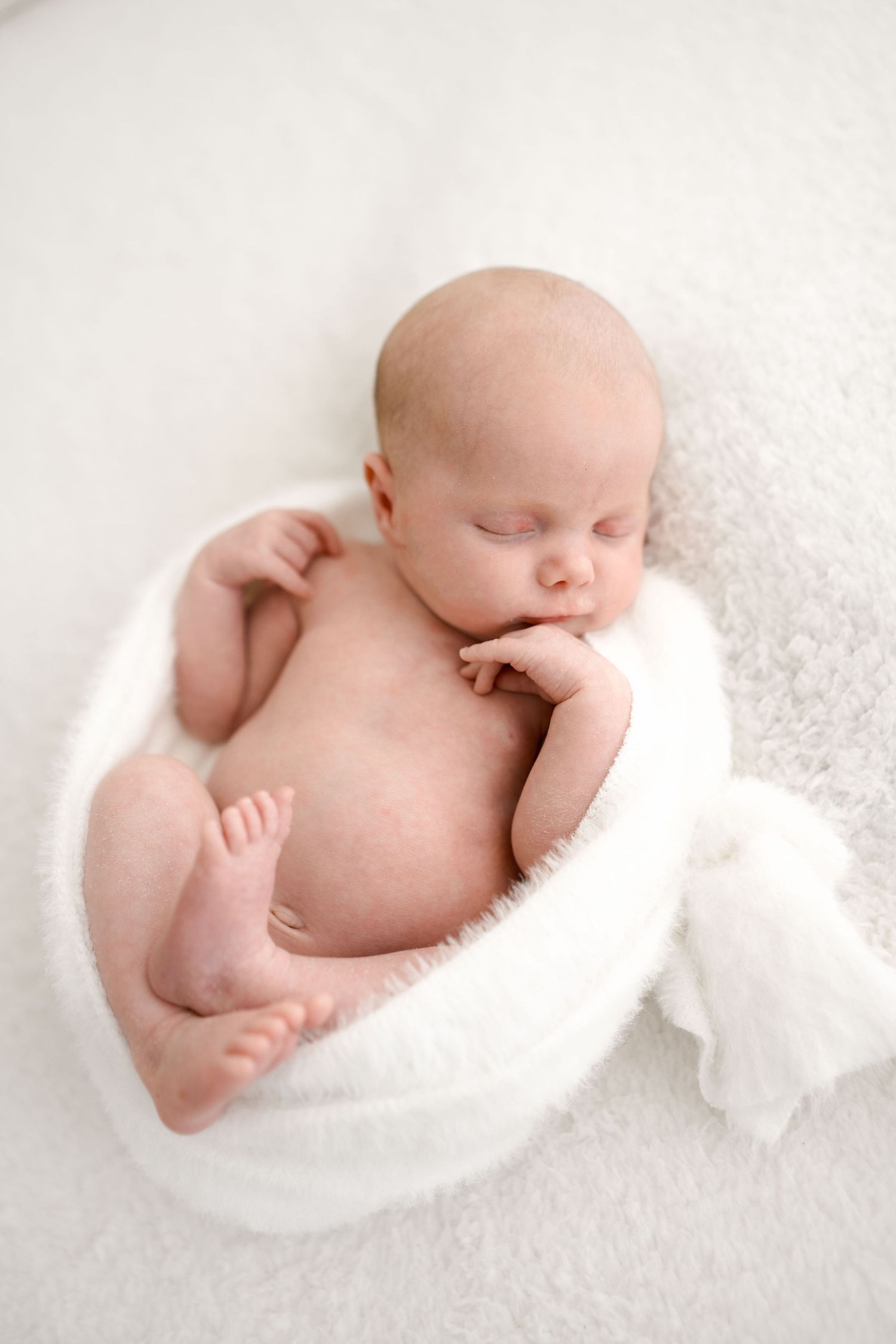 Peaceful newborn baby sleeping curled up in a white fluffy bowl-shaped prop on a white blanket.