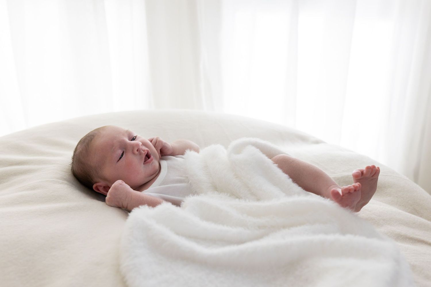Newborn baby lying on a white fluffy blanket in soft natural light, wrapped in a white swaddle.