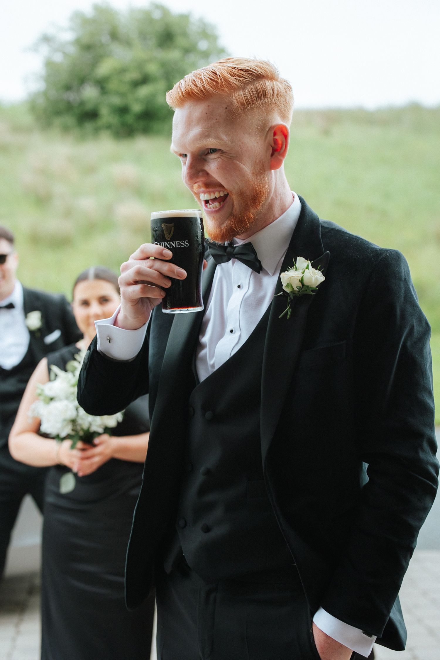 Smiling groom in black tuxedo with boutonniere drinking Guinness pint at outdoor wedding celebration.