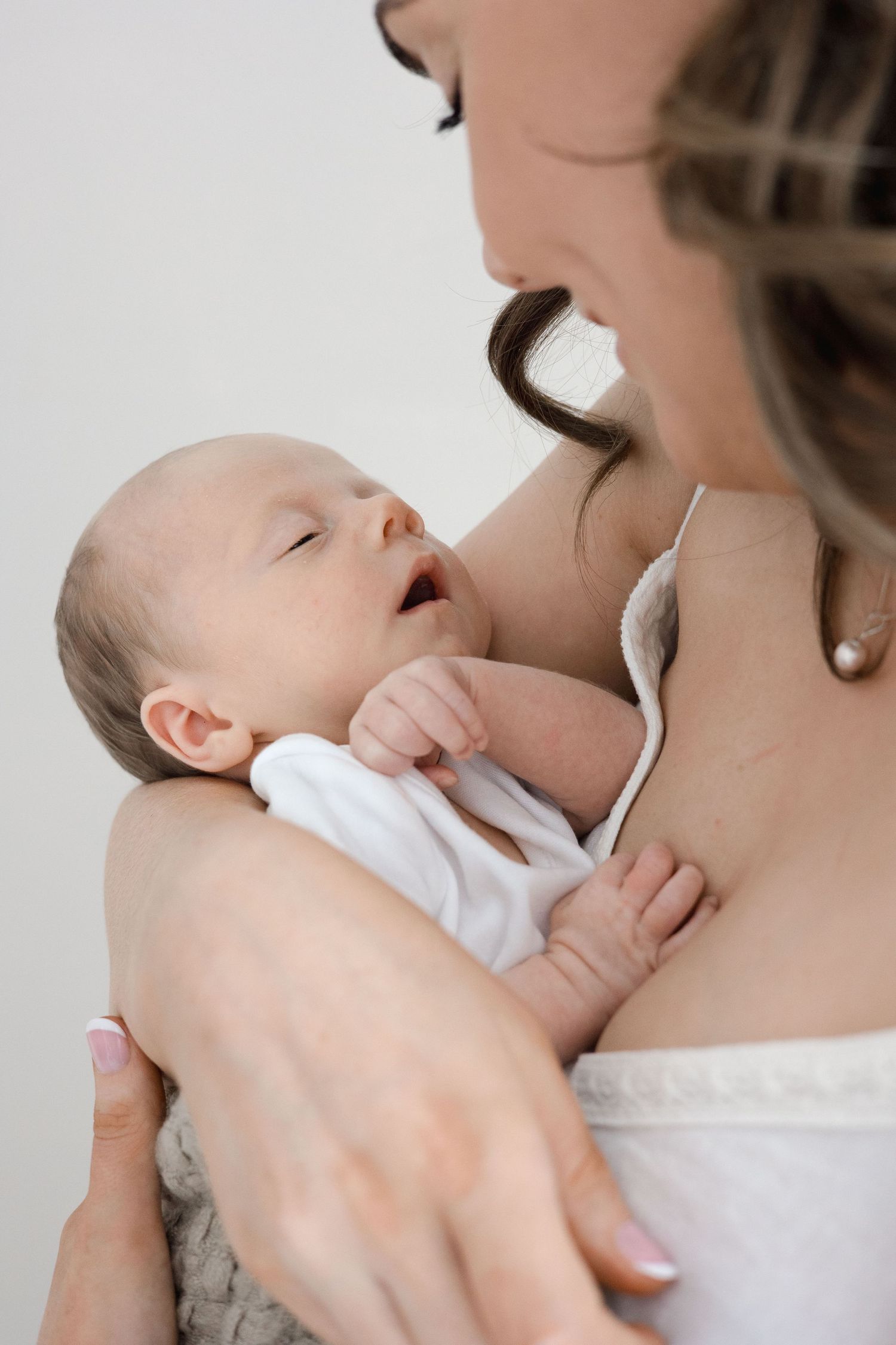 A mother cradles her sleeping newborn baby dressed in white against her chest in a bright studio.