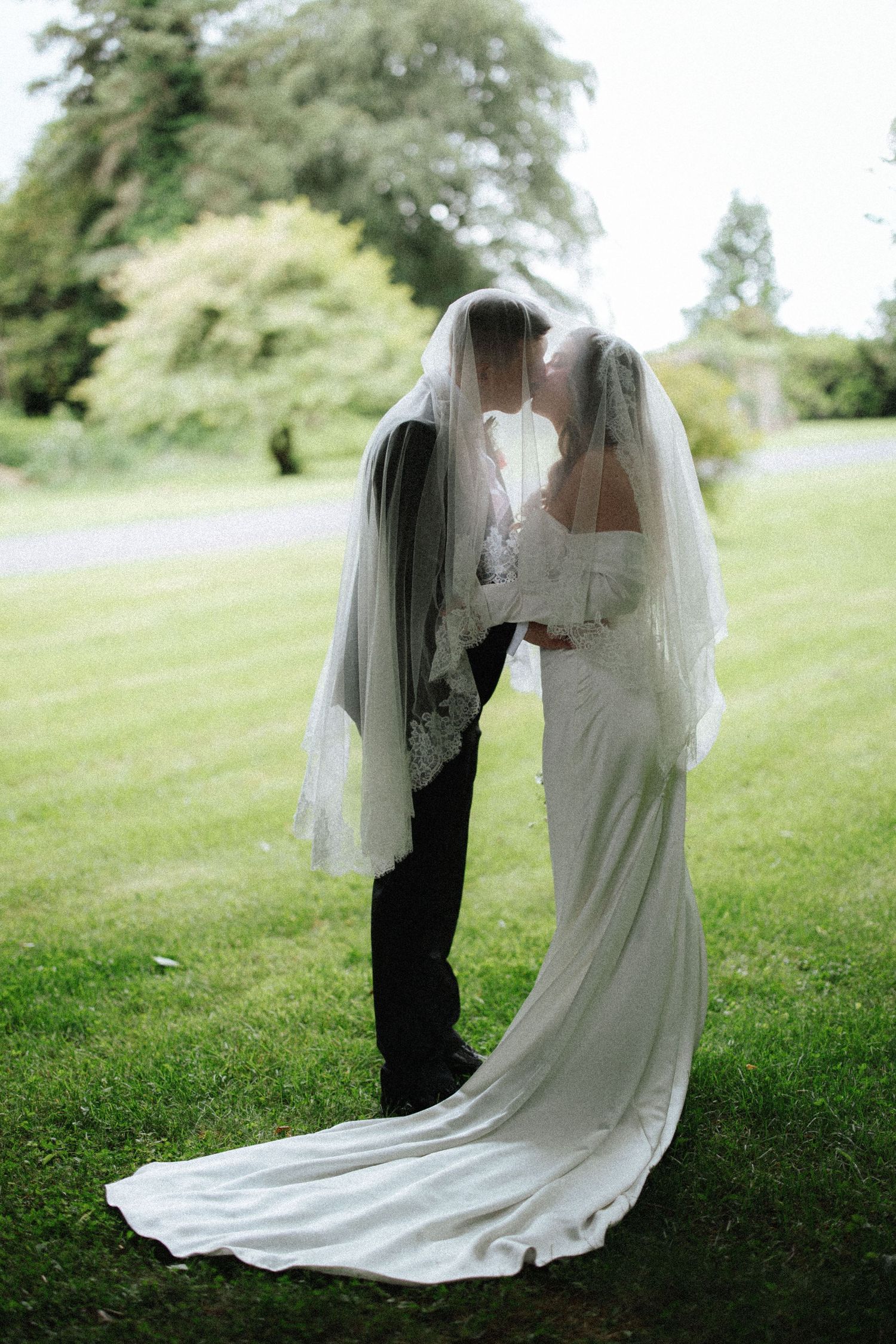 Bride and groom sharing a kiss under a wedding veil on a lush green lawn.