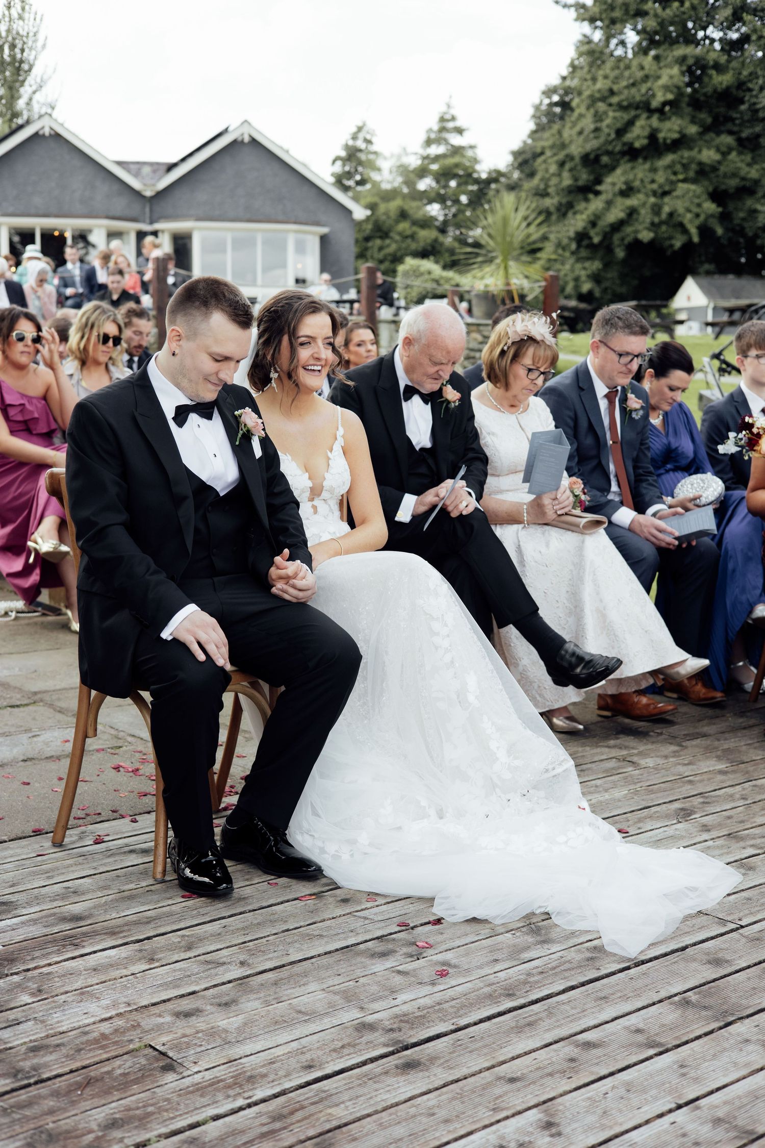 Bride in white gown and groom in tuxedo seated together laughing during their outdoor wedding ceremony.