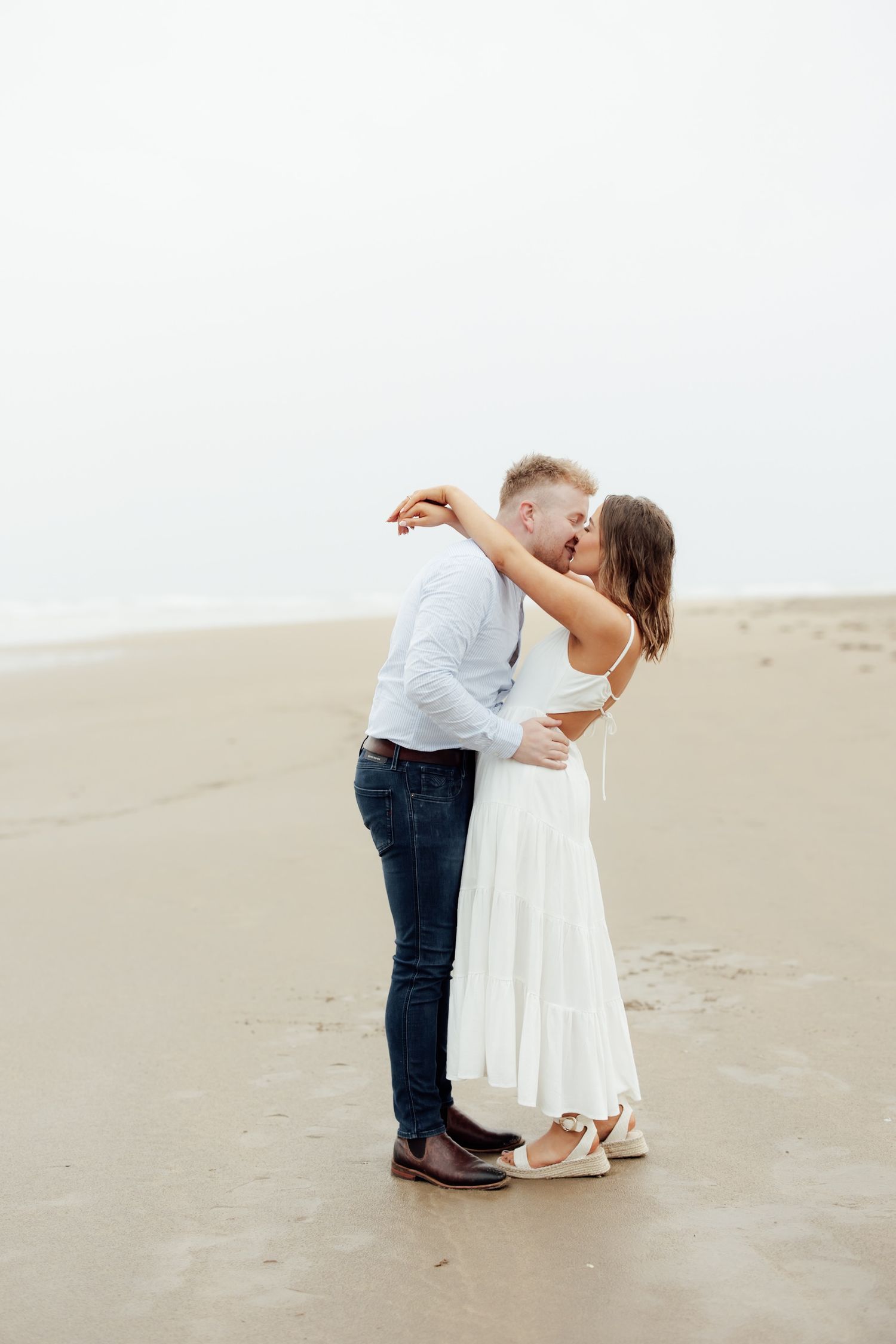 Couple kissing on a sandy beach, woman in white dress and man in blue jeans and white shirt.