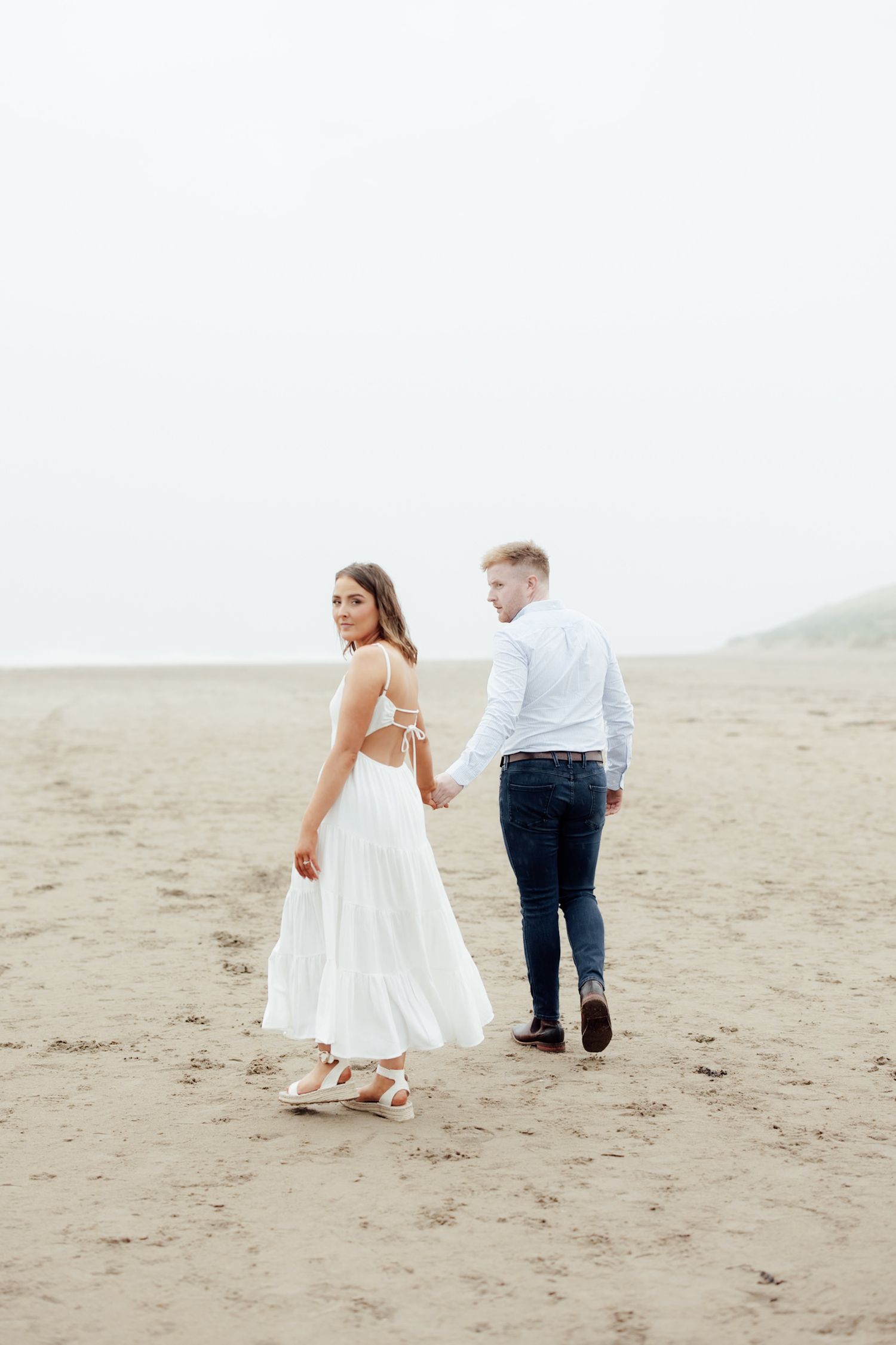 Couple walking hand in hand on a misty beach, woman in white dress glancing back, man in white shirt and jeans.