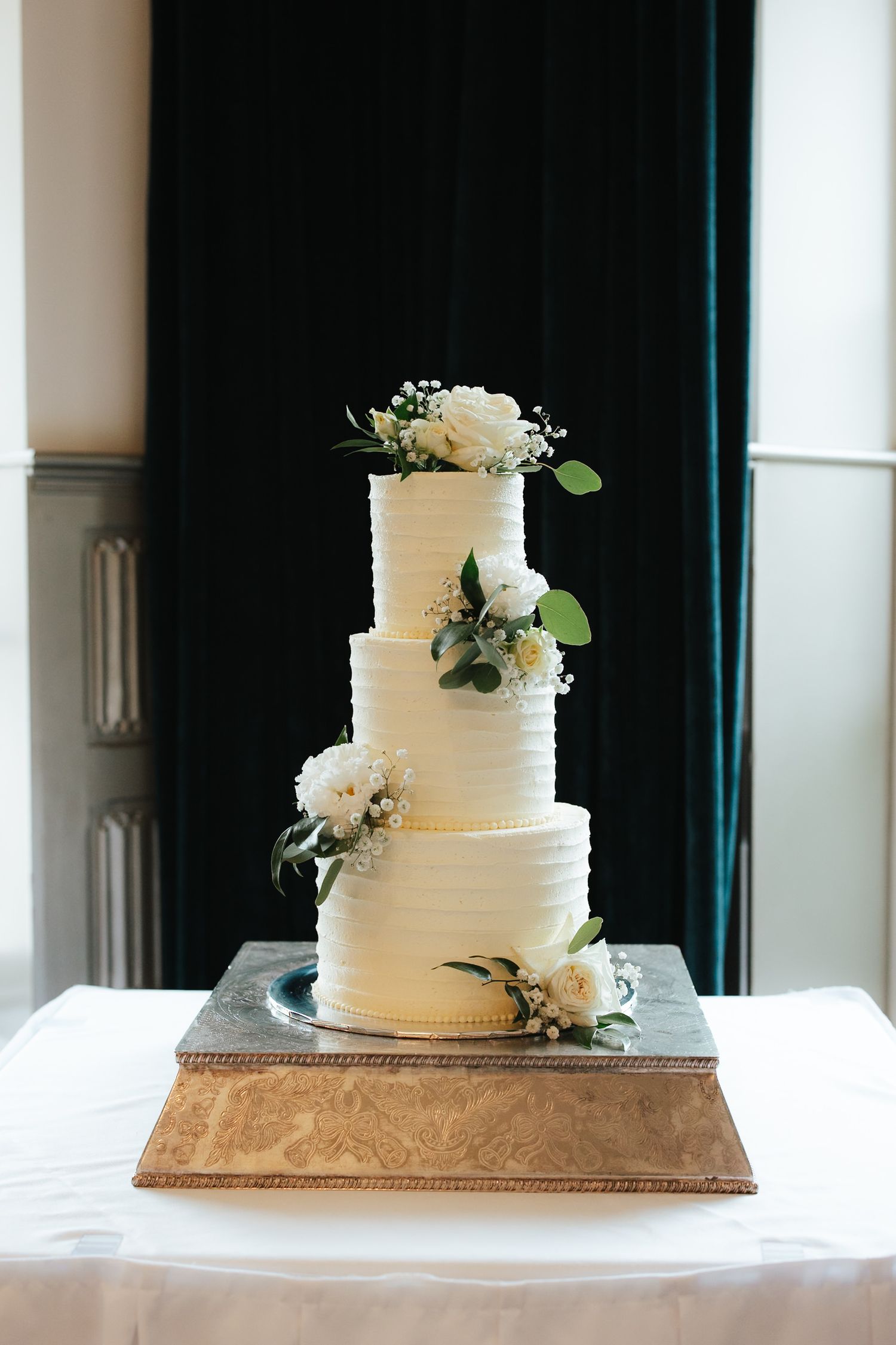 Four-tier white wedding cake with textured buttercream frosting and fresh white flowers on a silver stand.
