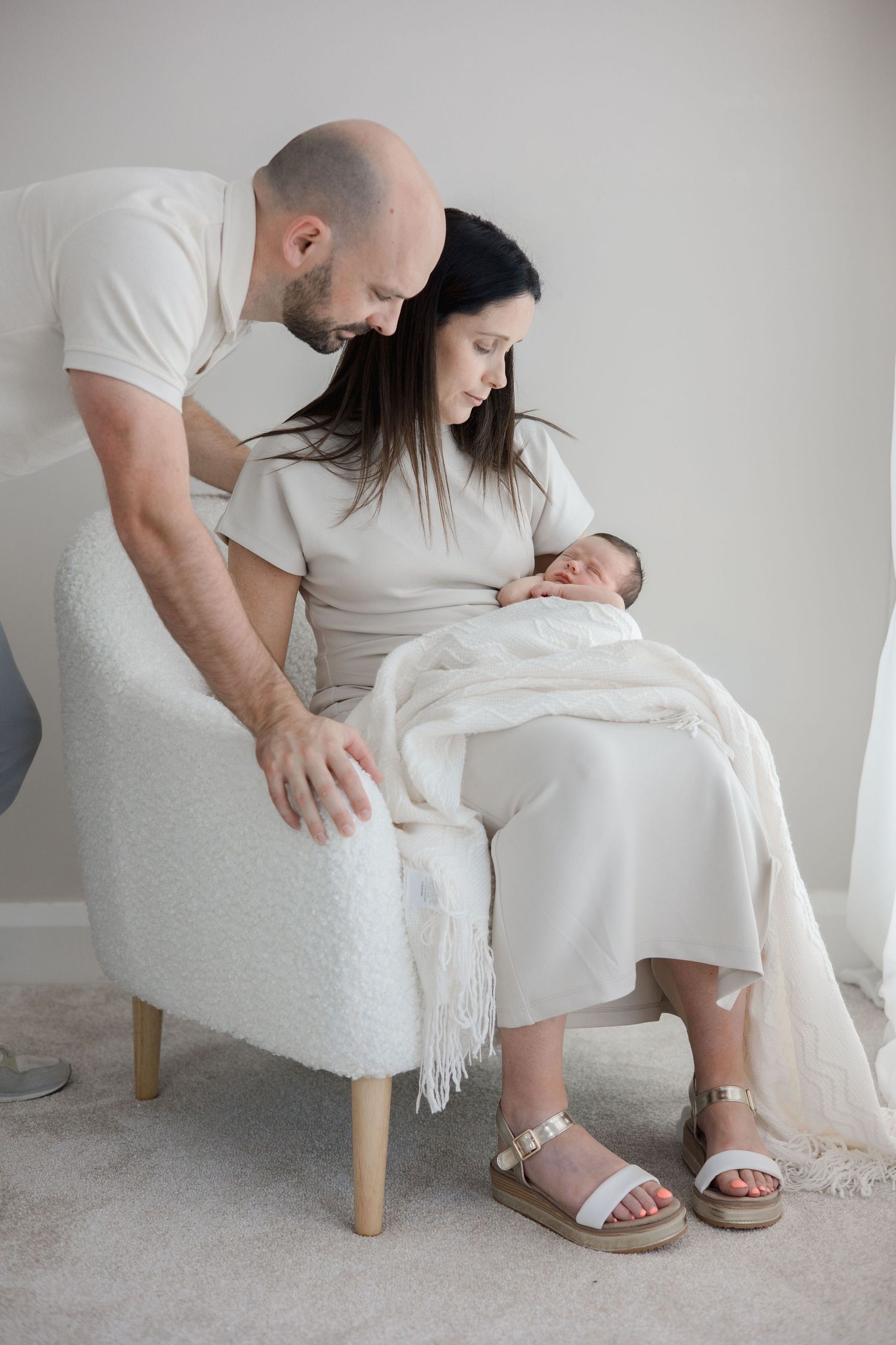 New parents lovingly admire their sleeping swaddled newborn as mother sits in white boucle armchair.