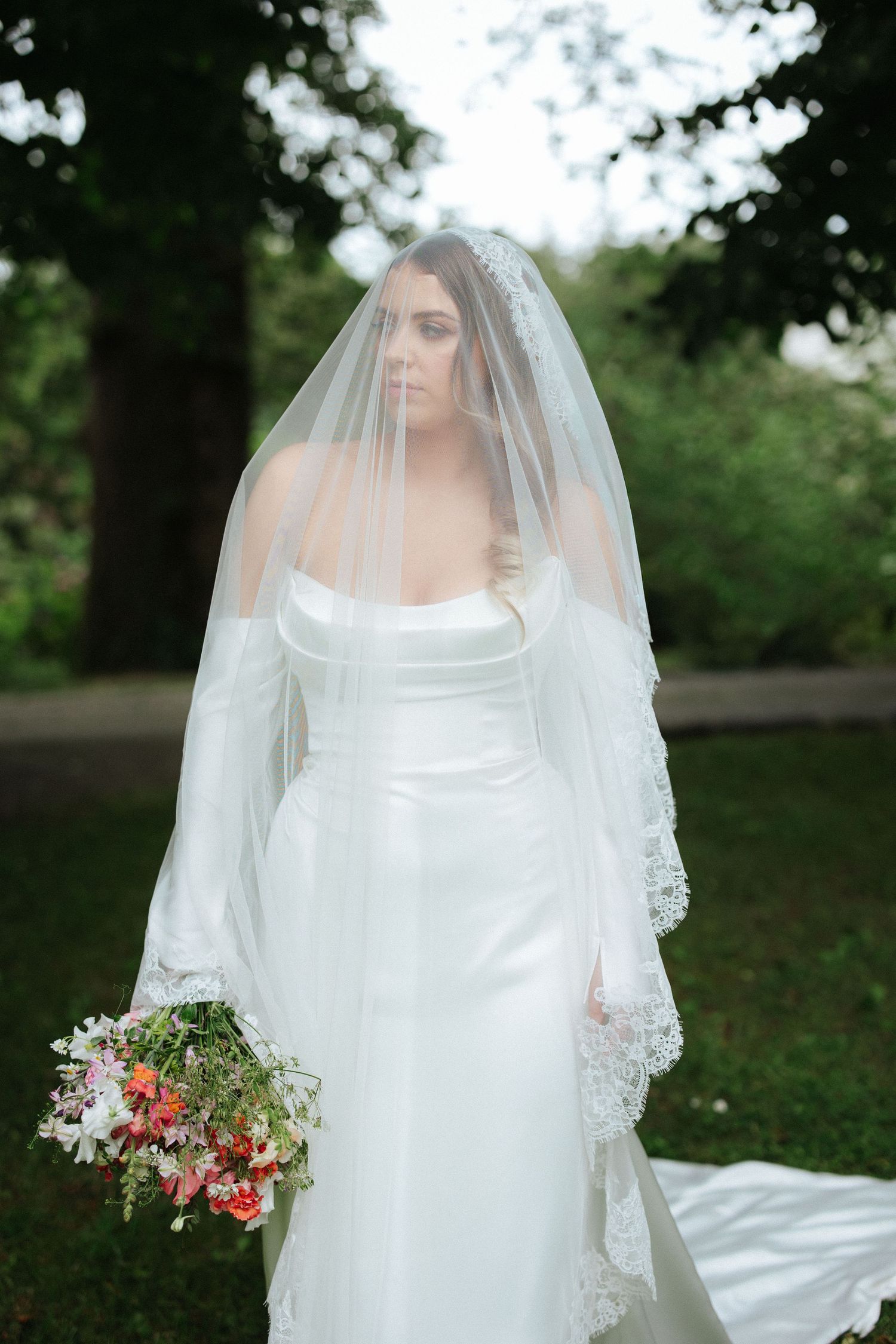 Bride in white satin gown and lace veil holding colorful wildflower bouquet outdoors.