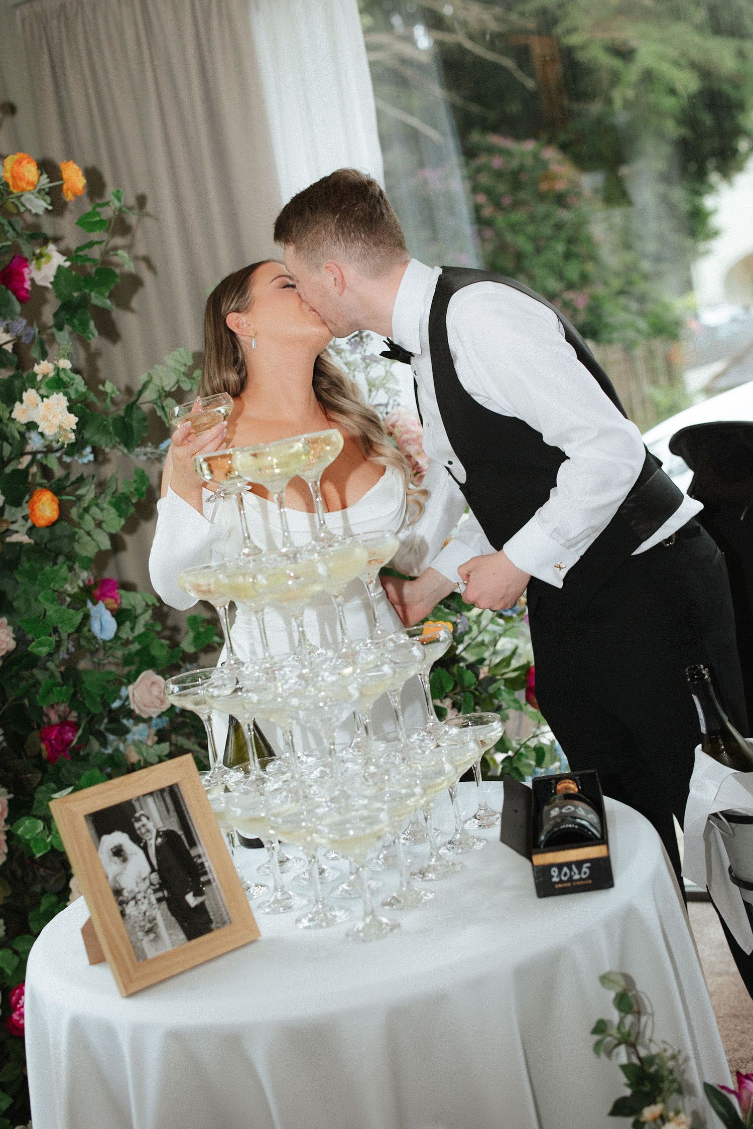 Bride and groom sharing a kiss beside a champagne tower at their wedding reception with floral arrangements.