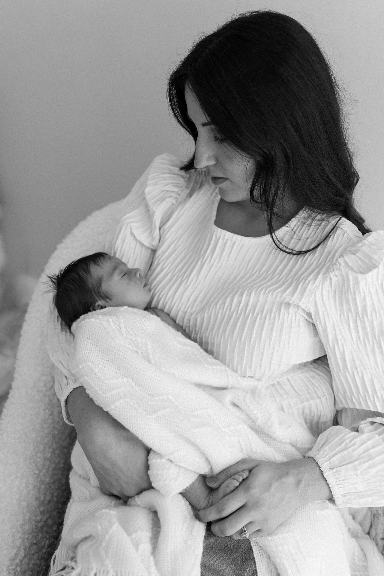 Black and white portrait of dark-haired mother tenderly holding her swaddled newborn baby.