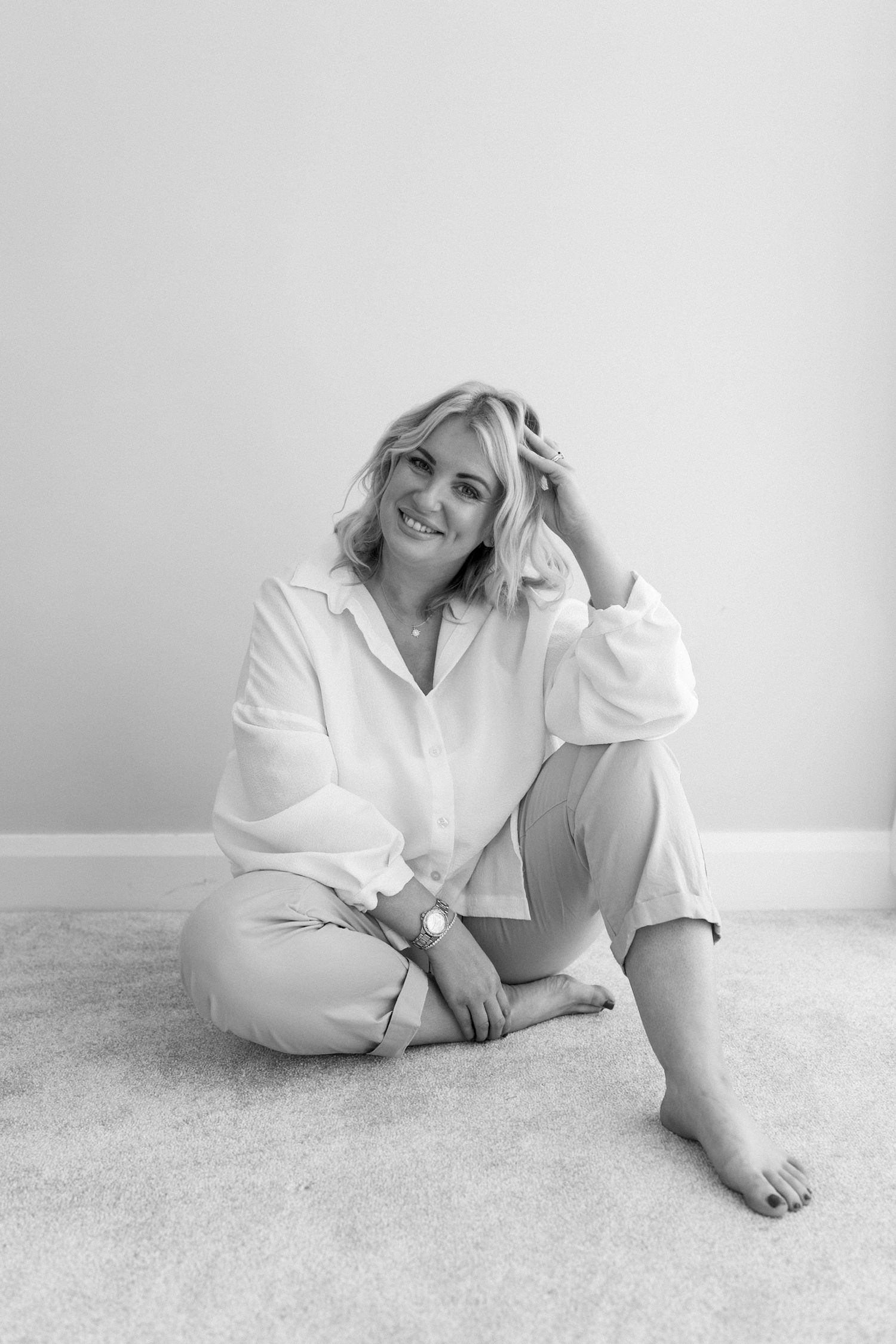 Smiling blonde woman in white shirt seated on floor in black and white lifestyle portrait.