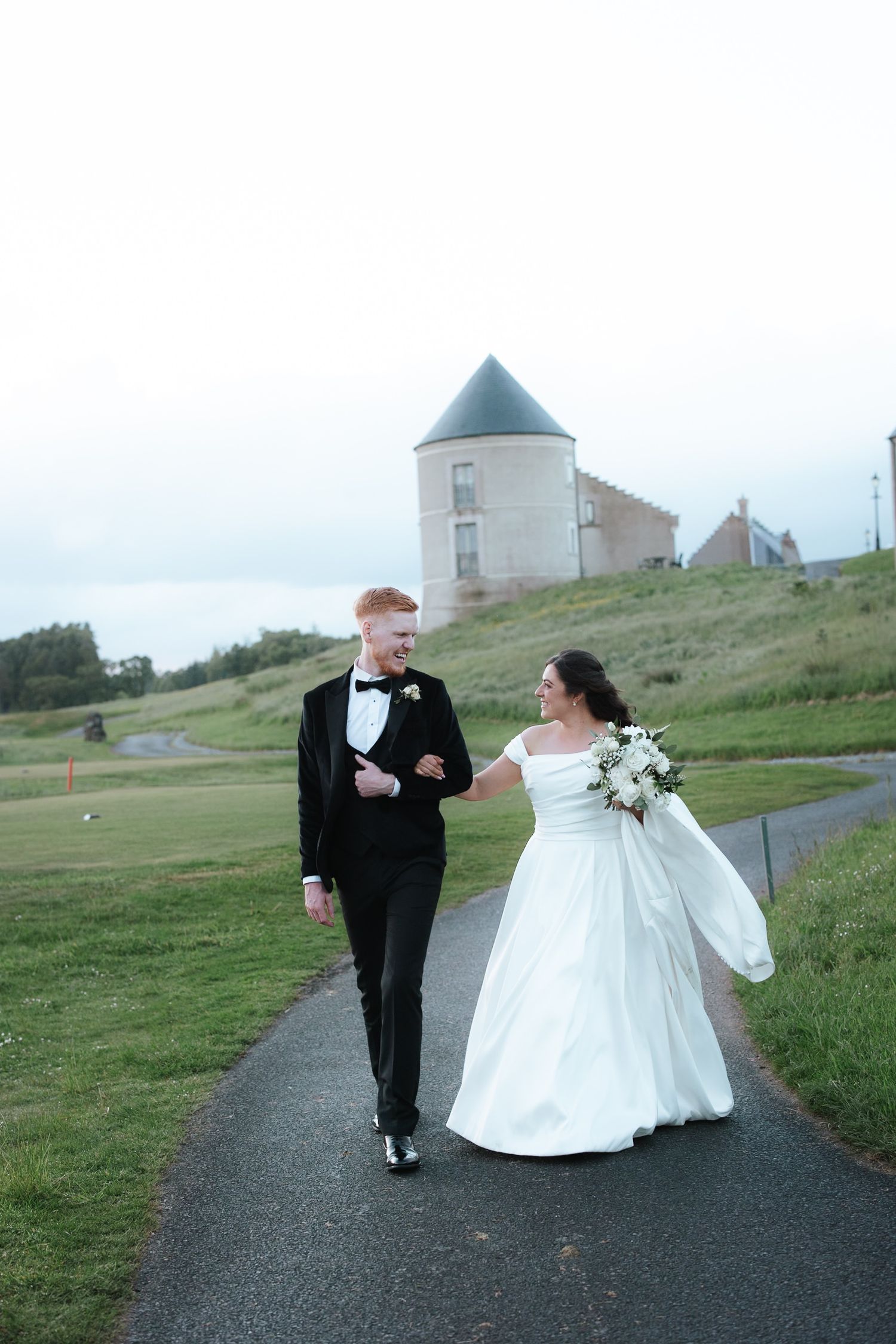 Bride and groom walk hand-in-hand on a path near a historic tower, bride holding white bouquet.