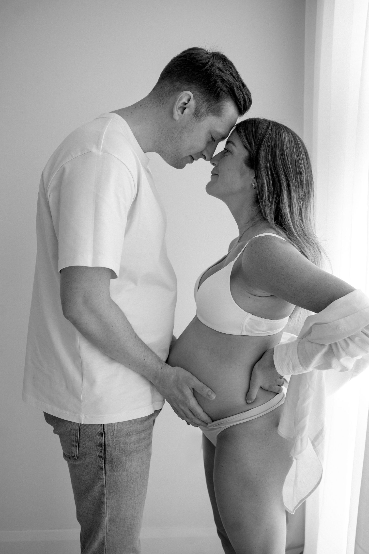 Black and white photo of expecting couple tenderly touching foreheads while holding pregnant belly.