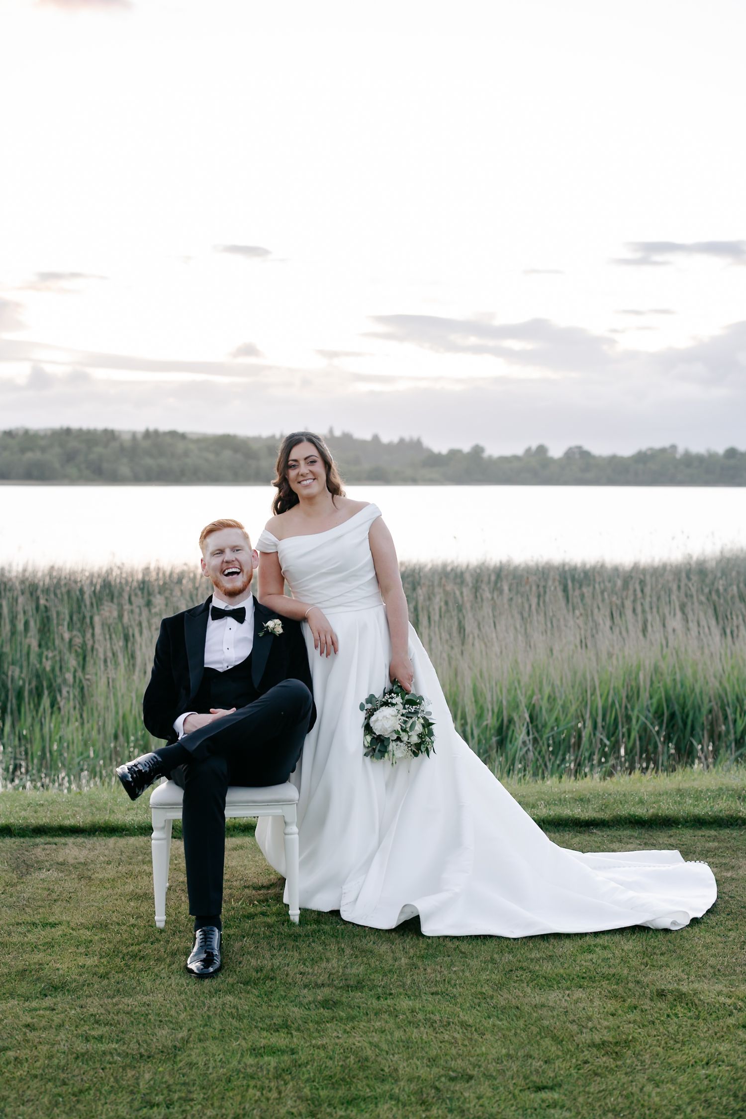 A smiling groom in a black tuxedo sits in a chair outdoors while his bride in a white gown stands beside him near a lake.