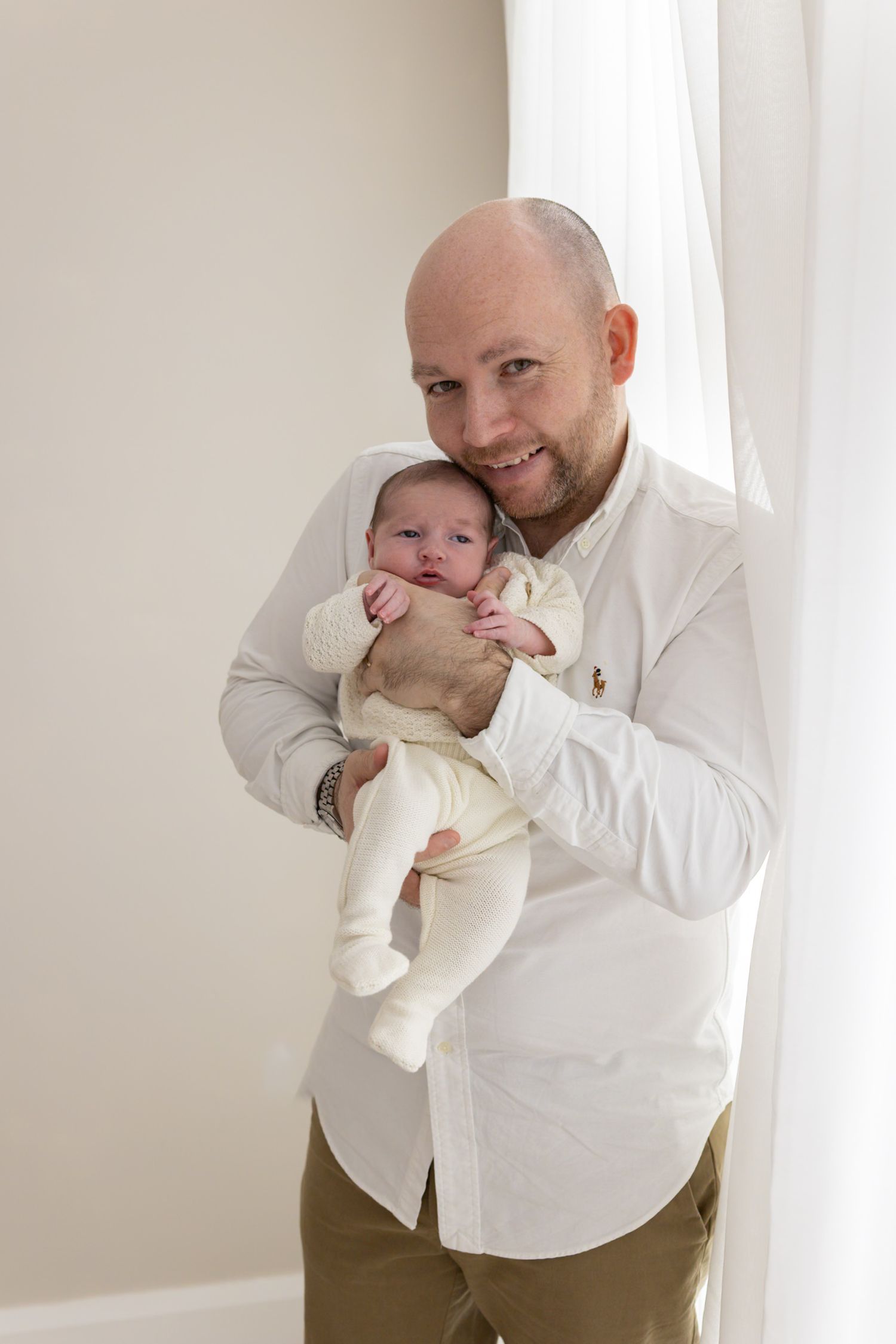 Proud father in white shirt smiling while holding his newborn baby by a window.