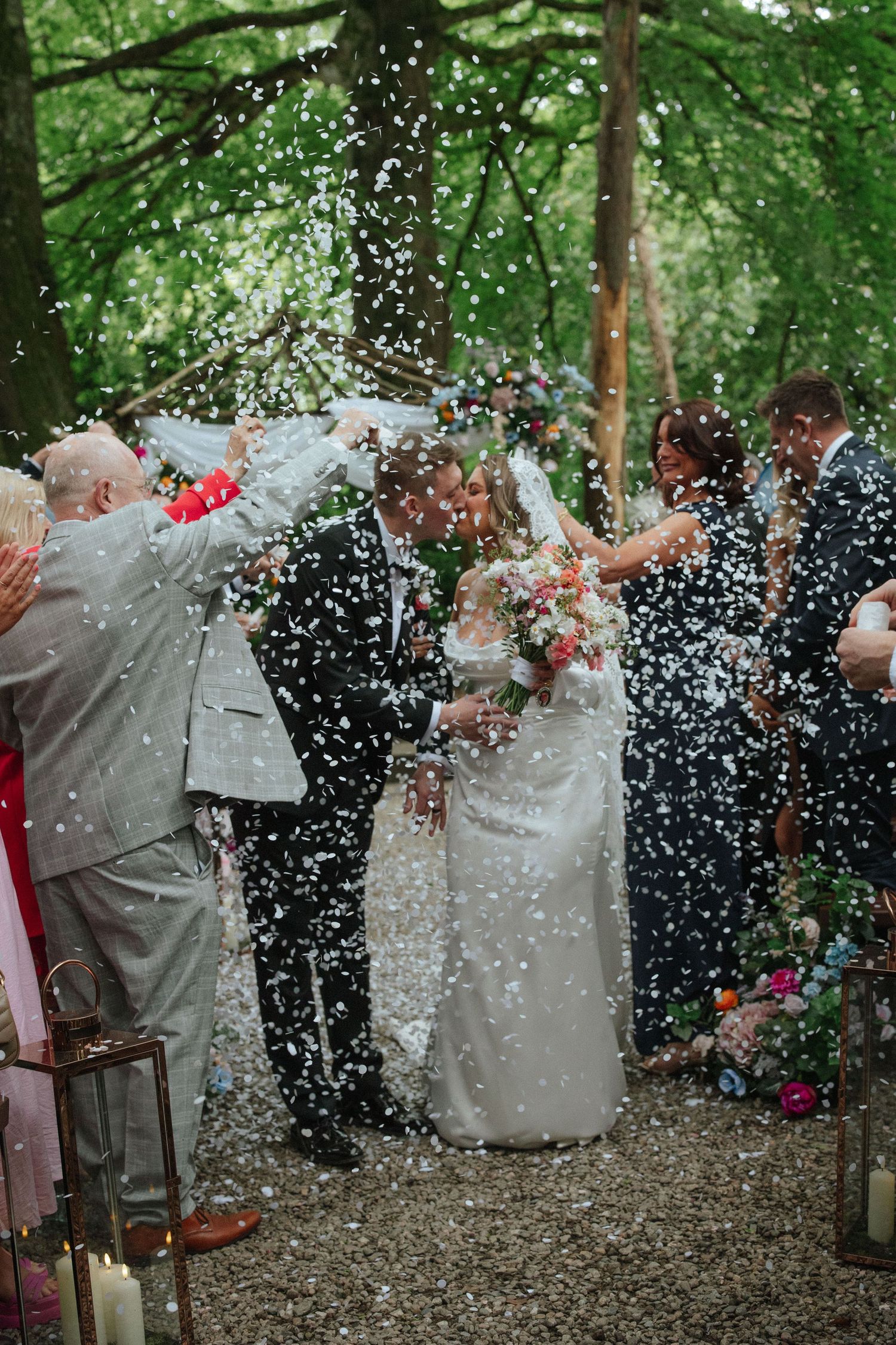Bride and groom kissing during confetti shower surrounded by wedding guests in a woodland ceremony setting.