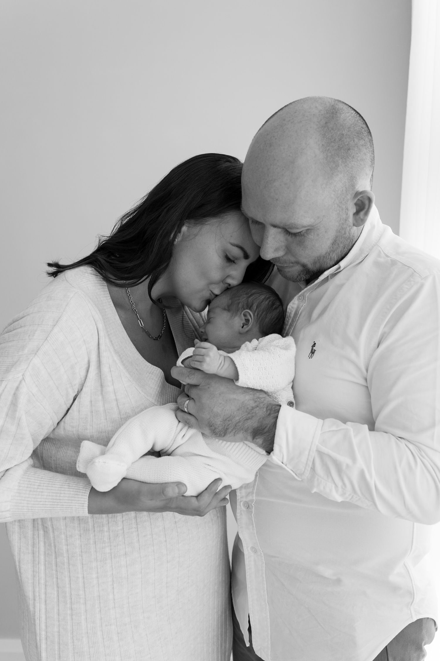 Black and white photo of parents tenderly kissing their newborn baby together.