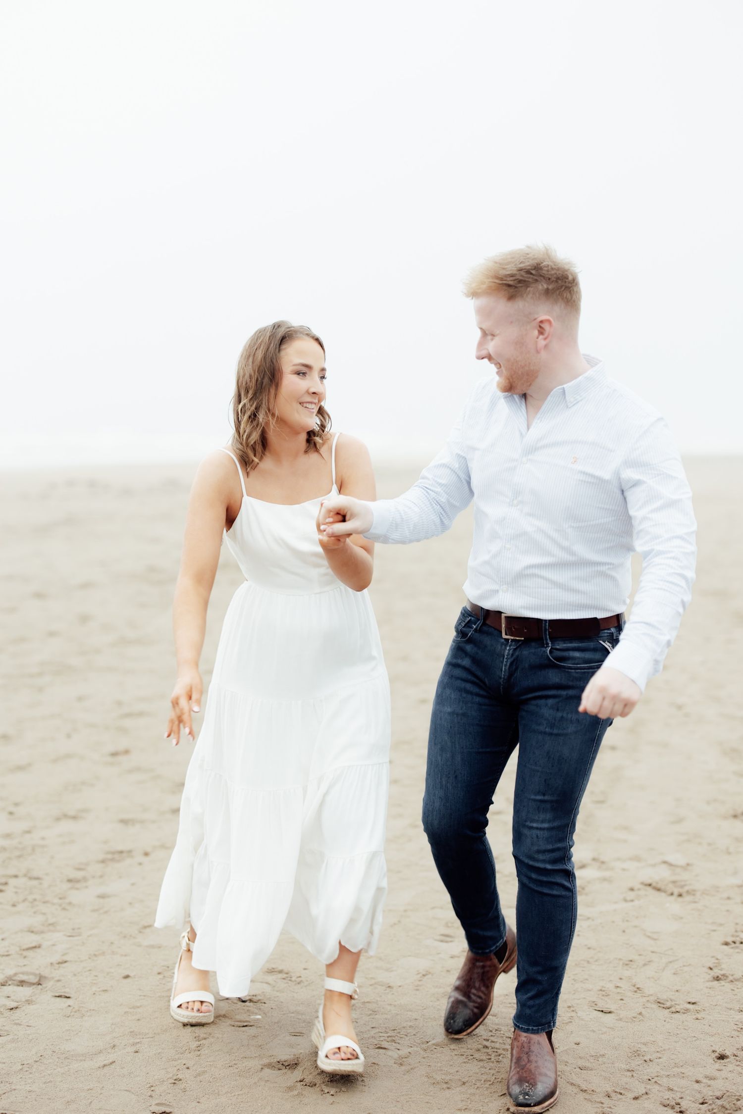 Couple walking hand in hand on a misty beach, woman in white dress, man in jeans and white shirt.