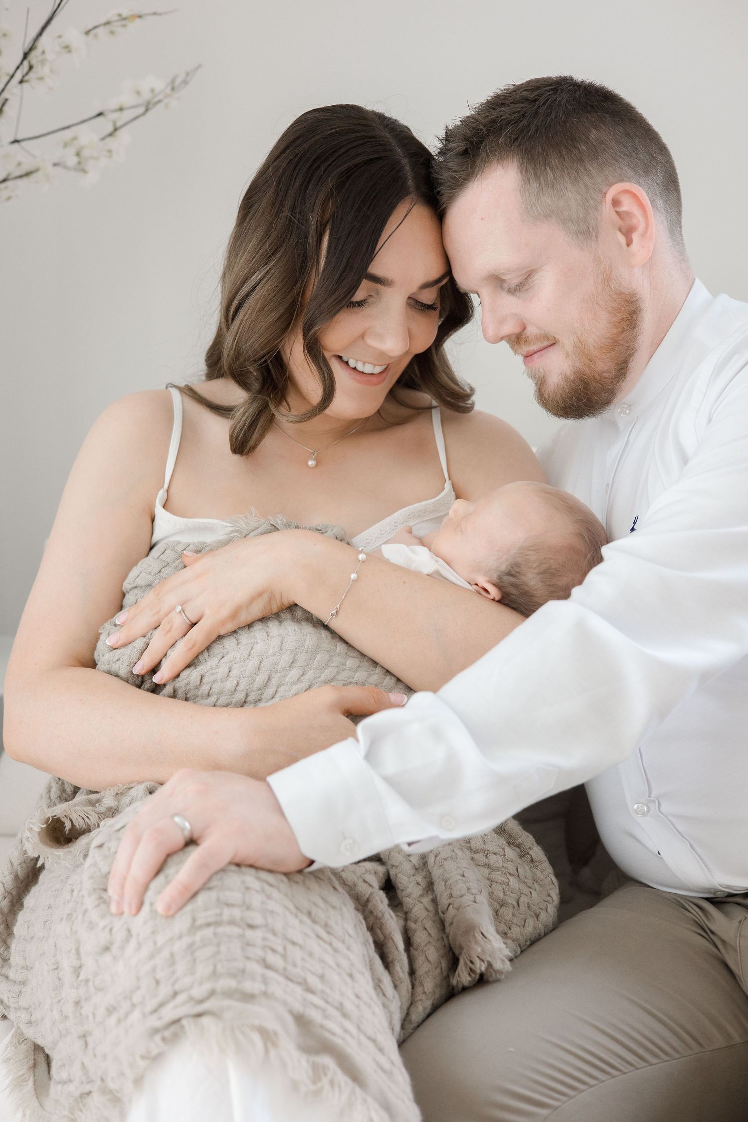 Smiling parents lean together over their newborn baby wrapped in a cozy gray blanket in a bright studio.