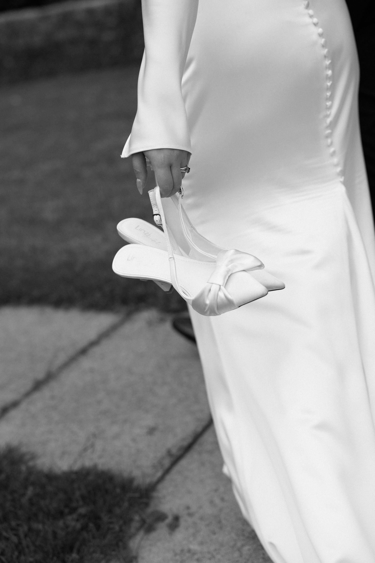 Bride in white wedding dress holding satin bridal shoes, black and white photography.
