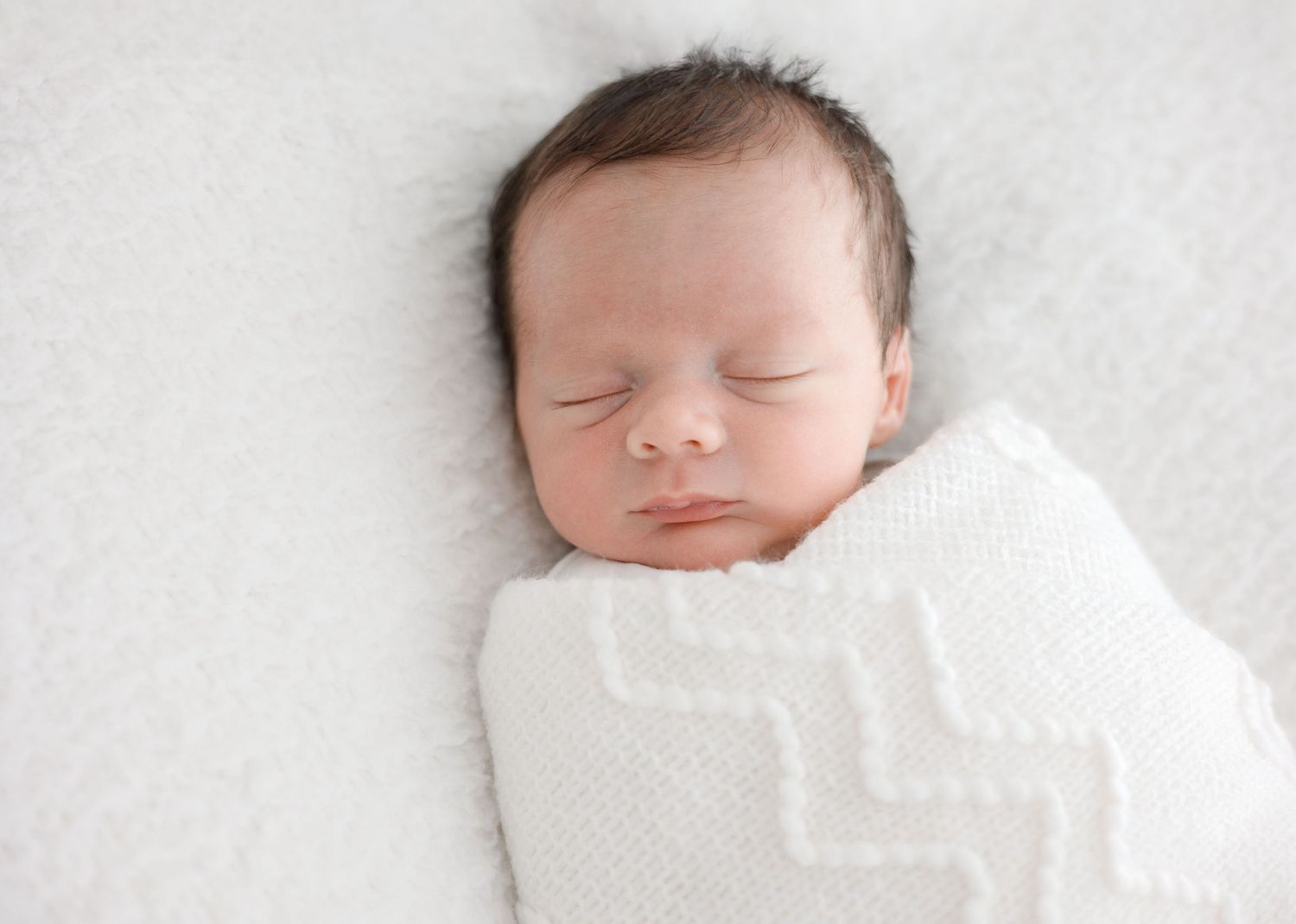 Peaceful newborn baby swaddled in white knit blanket sleeping on soft white fluffy surface.