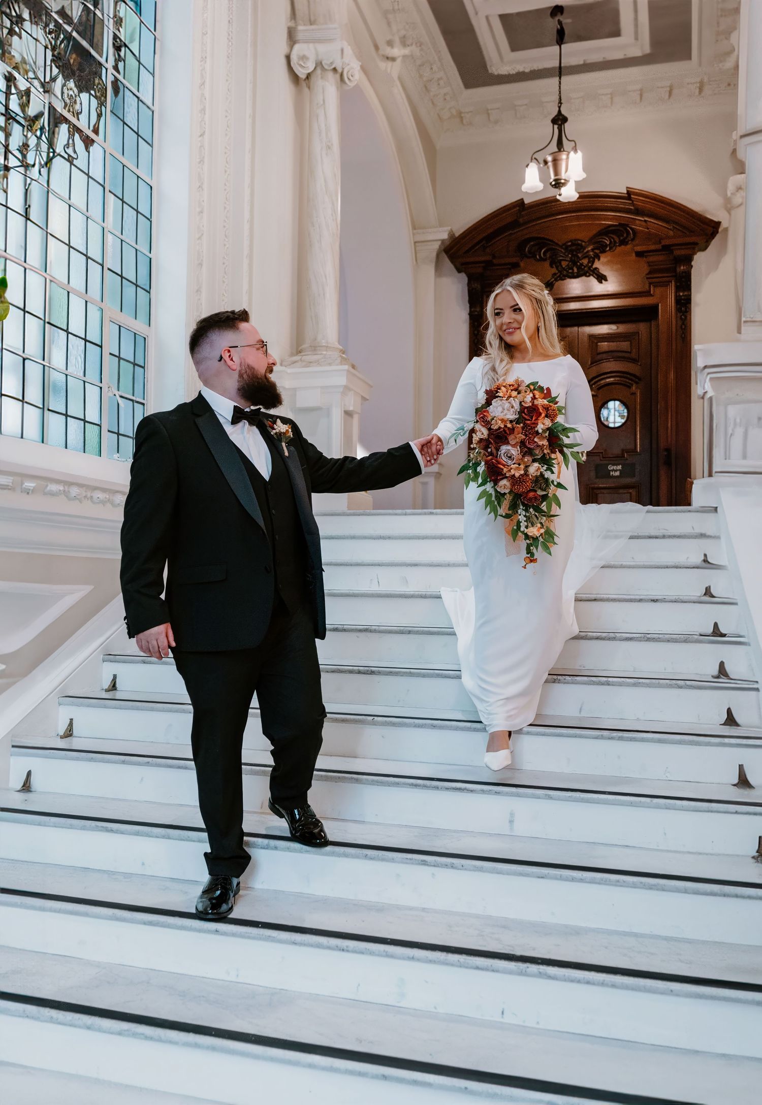 Elegant bride and bearded groom descend grand marble staircase in ornate wedding venue holding hands.