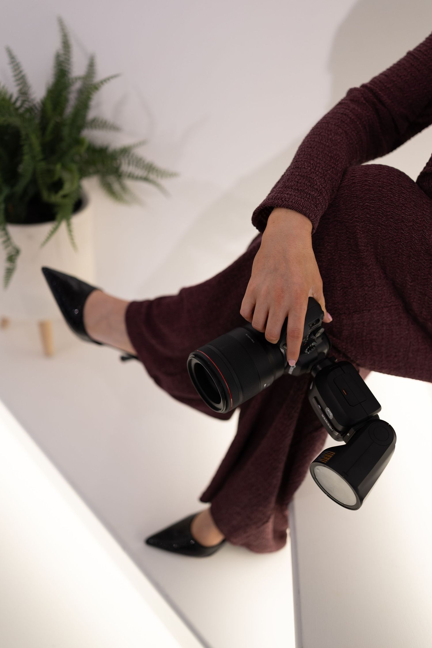 Close-up of hands holding professional camera with burgundy pants and pointed black heels.