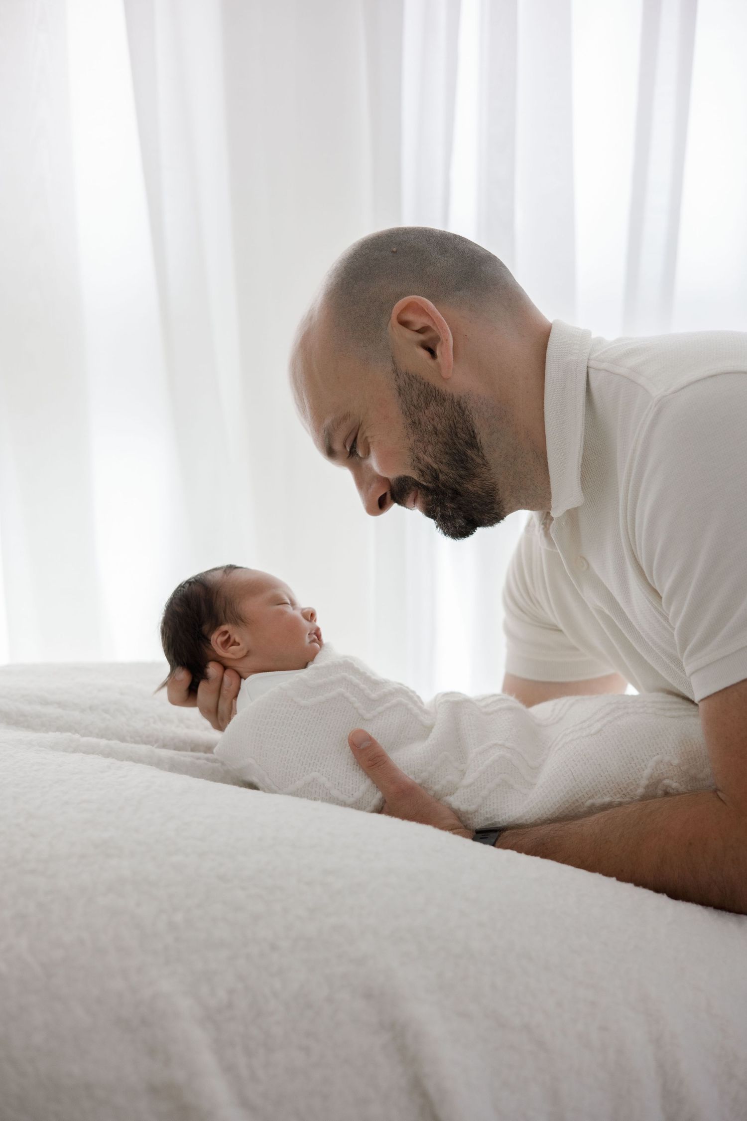 Loving bald father leans over white bed gazing warmly at his swaddled newborn baby in bright room.