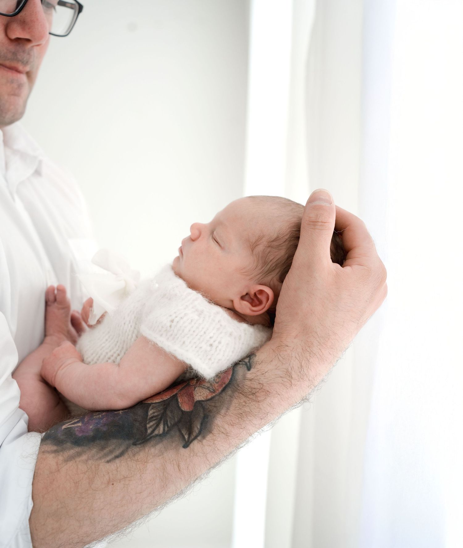 Tattooed father in white shirt gently cradling sleeping newborn baby in his arms.