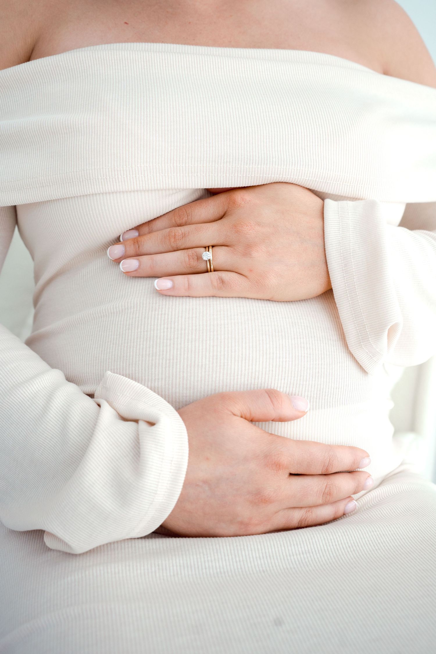 Close-up of pregnant woman in white off-shoulder dress gently cradling her baby bump.