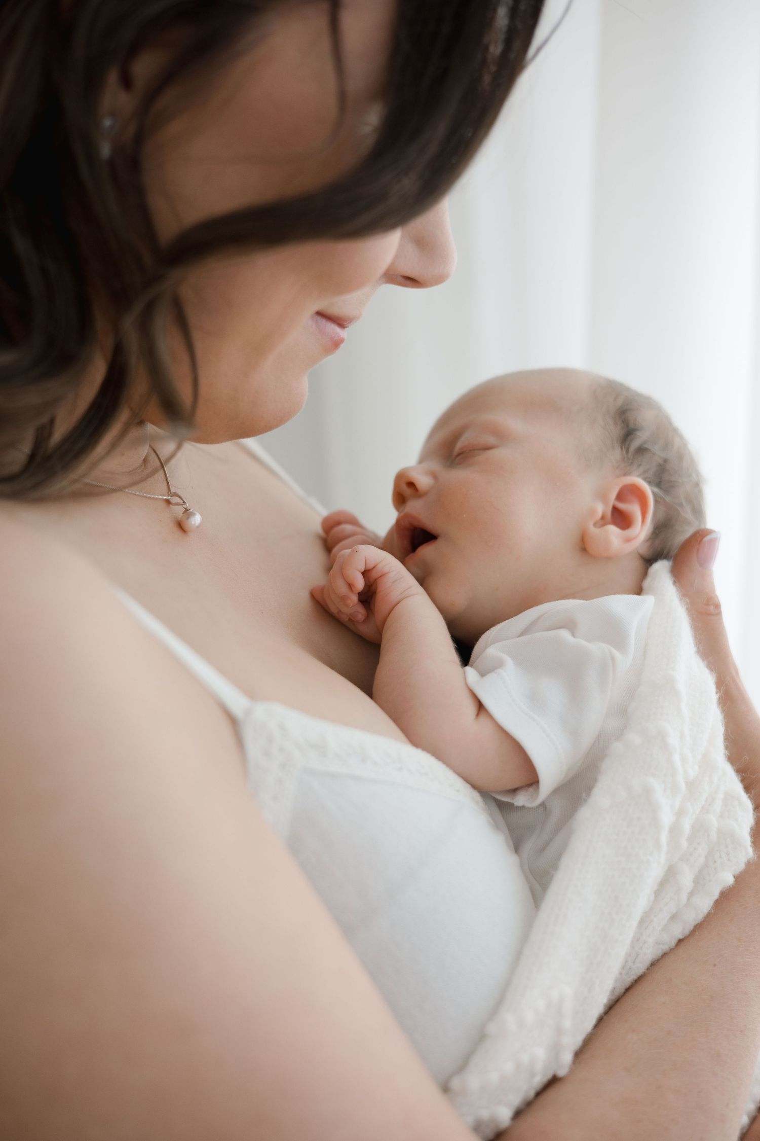 A mother gazes lovingly at her newborn baby cradled in her arms near a bright window.