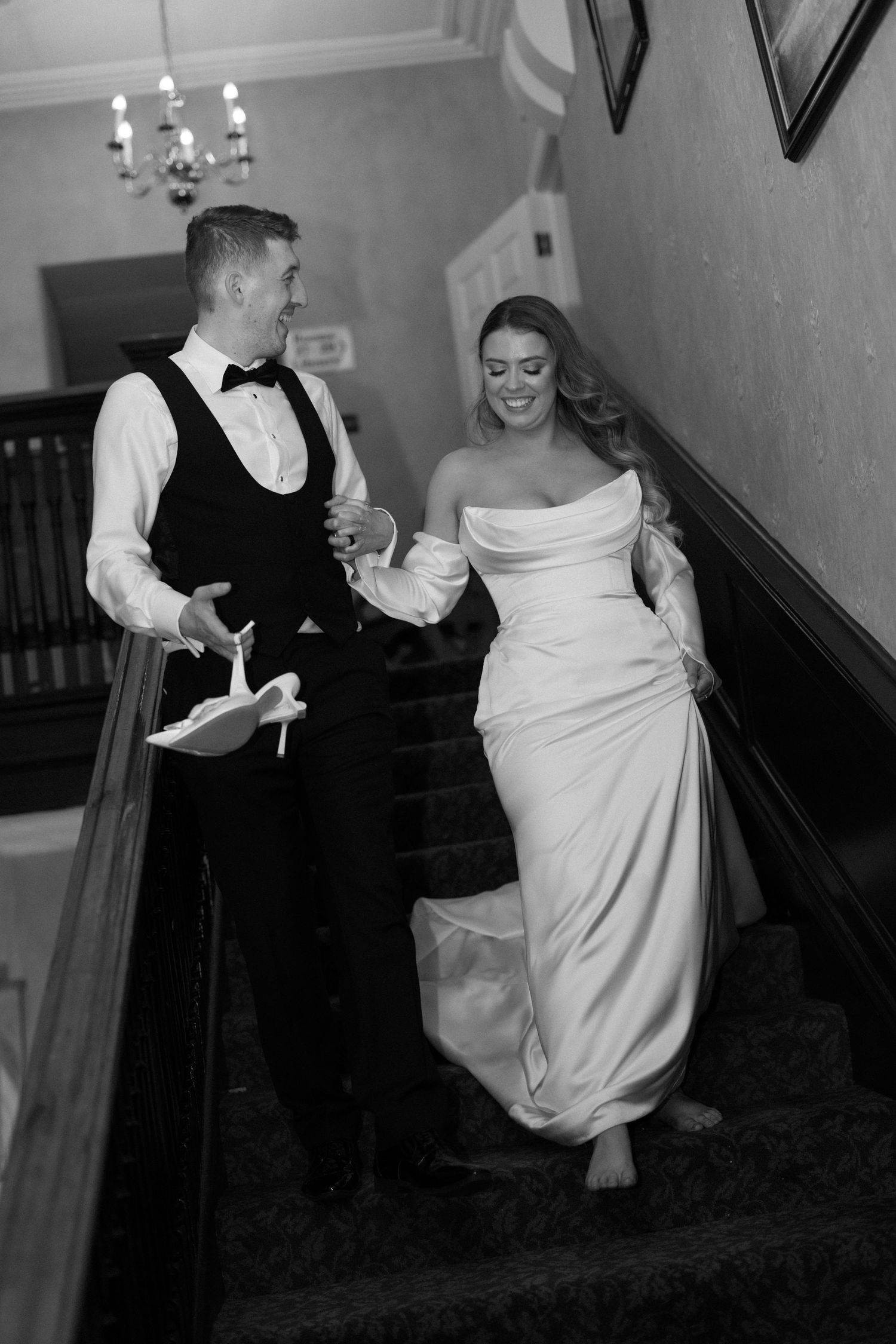 Black and white photo of a smiling bride and groom walking down a staircase, bride in strapless gown, groom in vest and bow tie.
