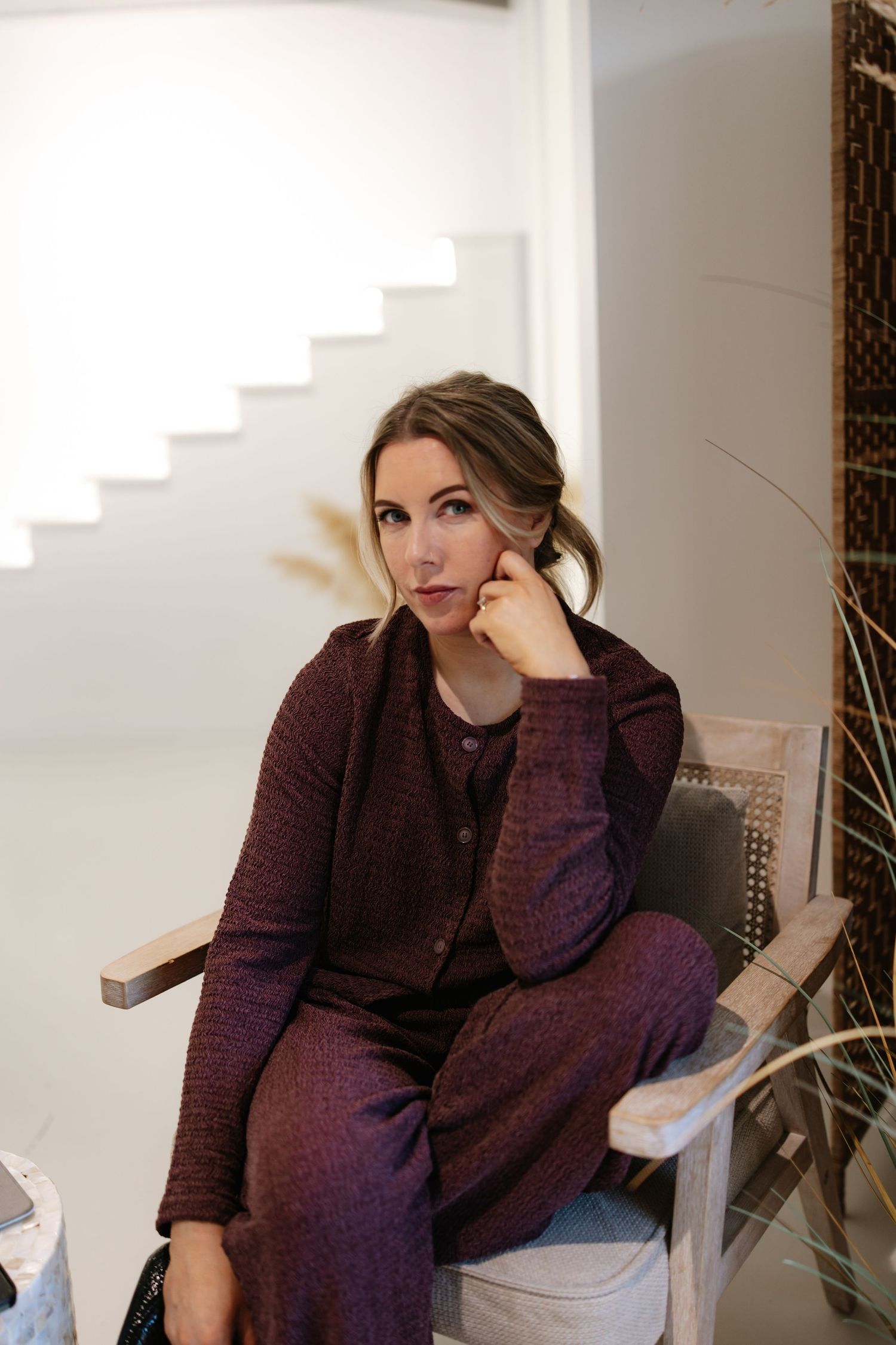 Woman in burgundy knit set sitting thoughtfully in chair in bright modern interior space.