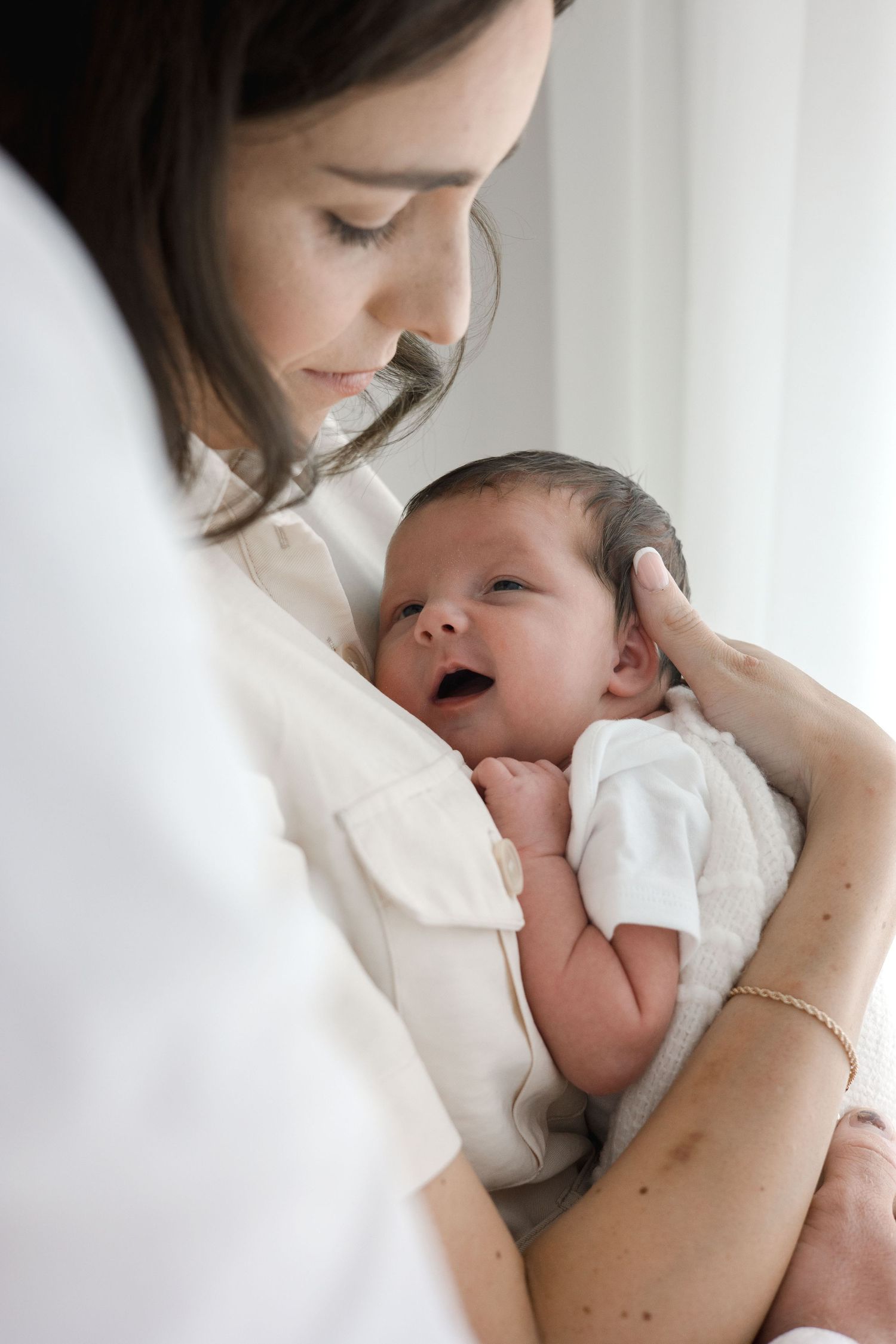 Mother in white shirt tenderly holds her smiling newborn baby against a bright window.