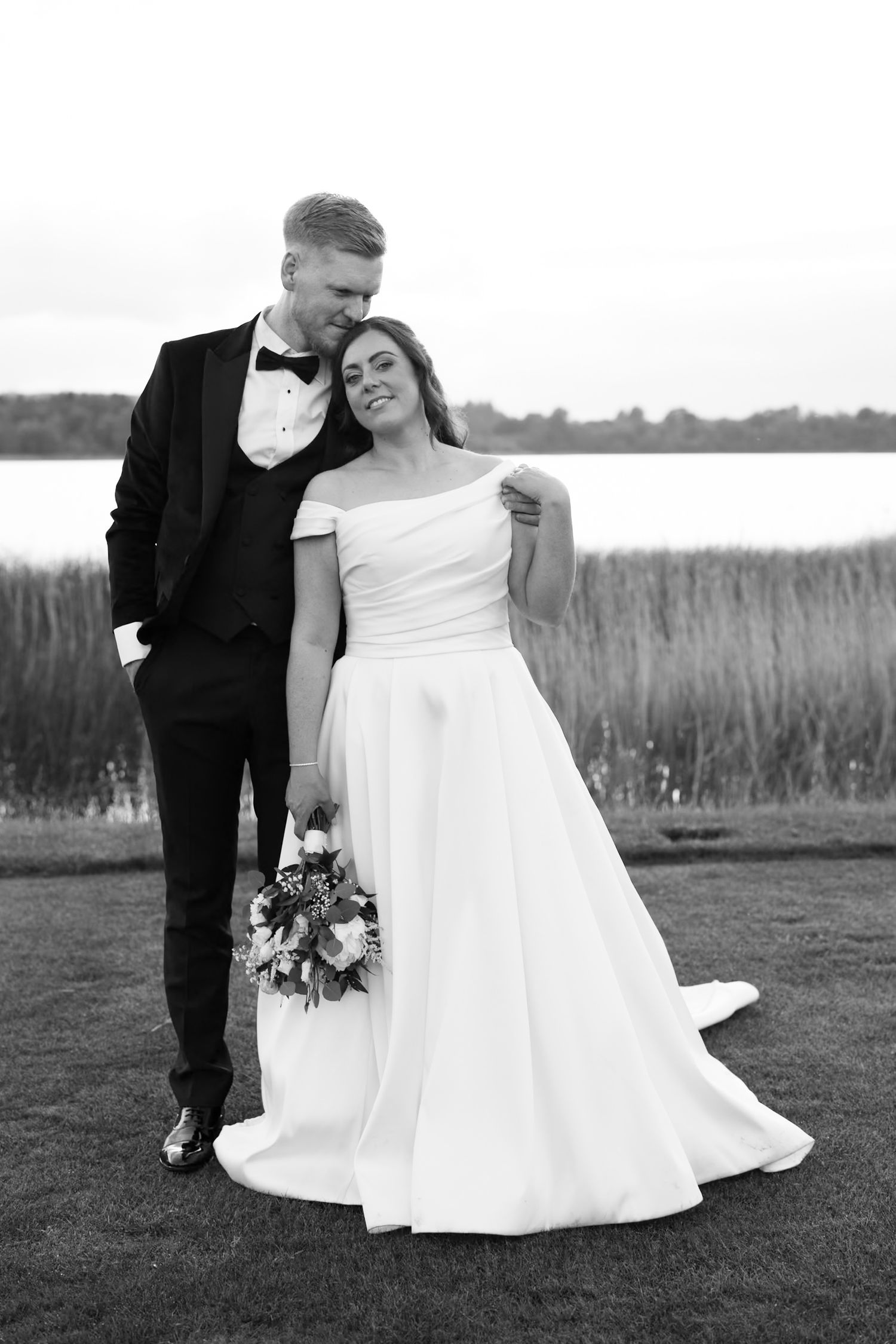 Black and white photo of a bride in white gown and groom in tuxedo posing together near a lake.