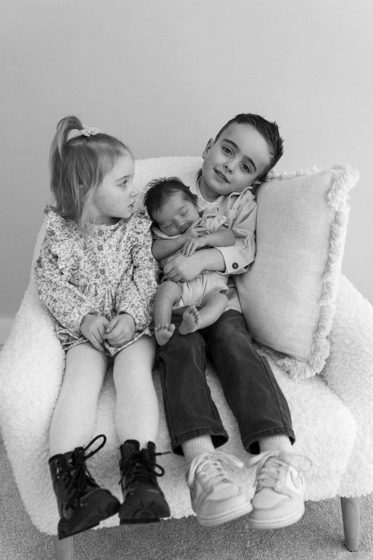 Black and white photo of two siblings sitting in a white chair holding their sleeping newborn baby.