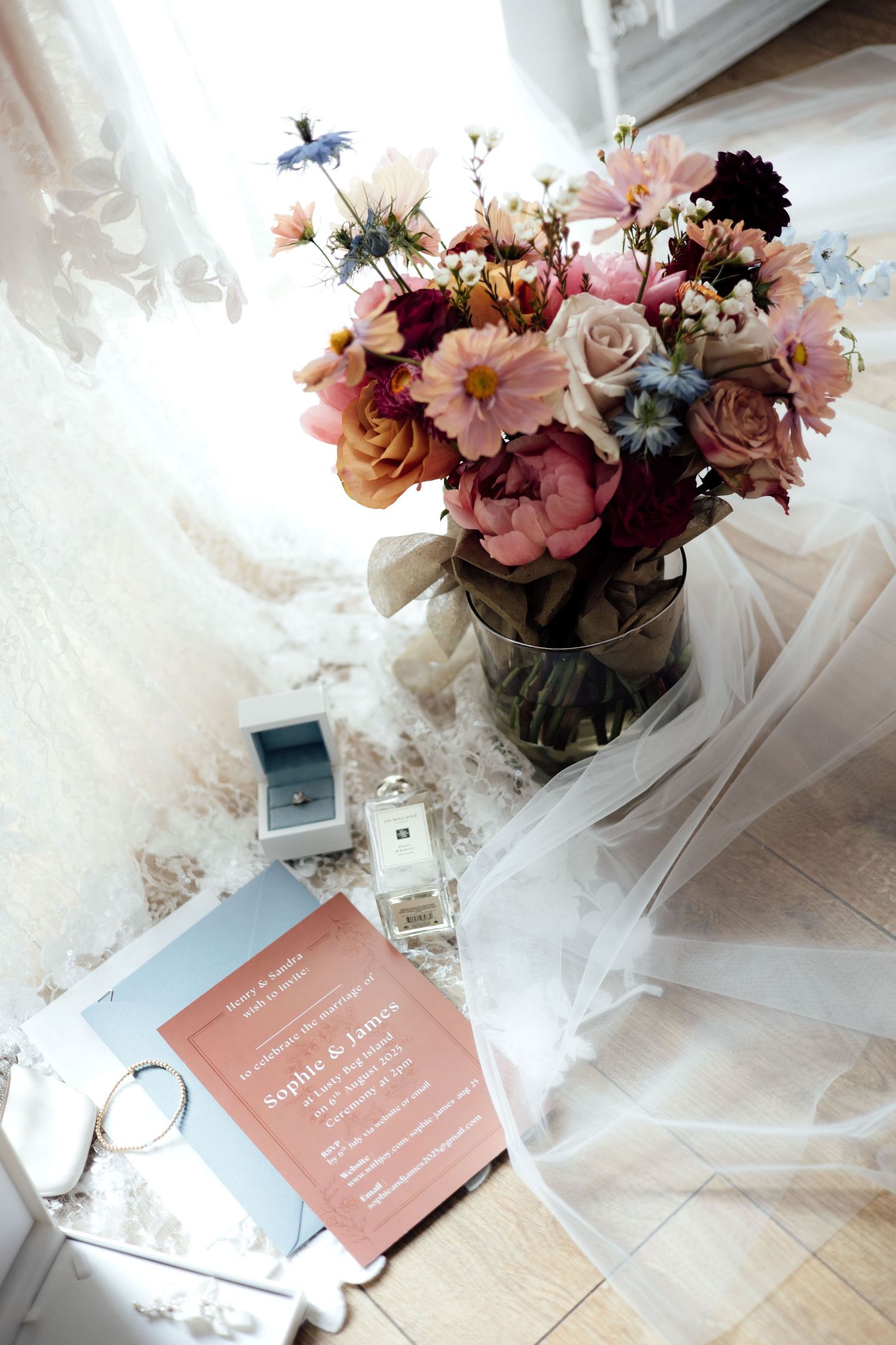 Colorful bridal bouquet with peonies and roses beside wedding invitation, perfume, and veil.