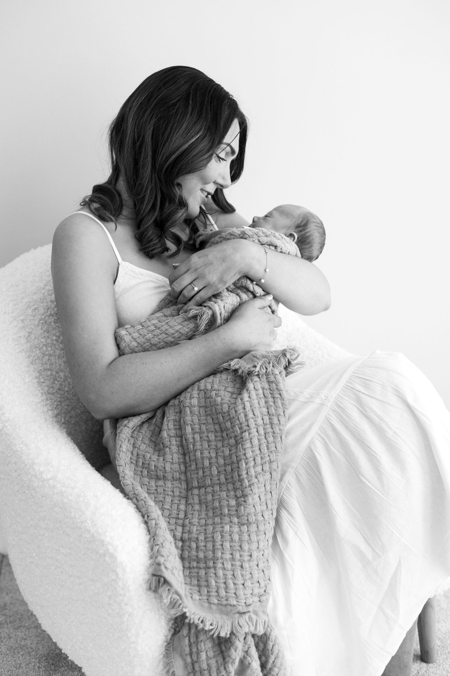 A smiling dark-haired mother sits in a white chair cradling her newborn in a textured blanket.