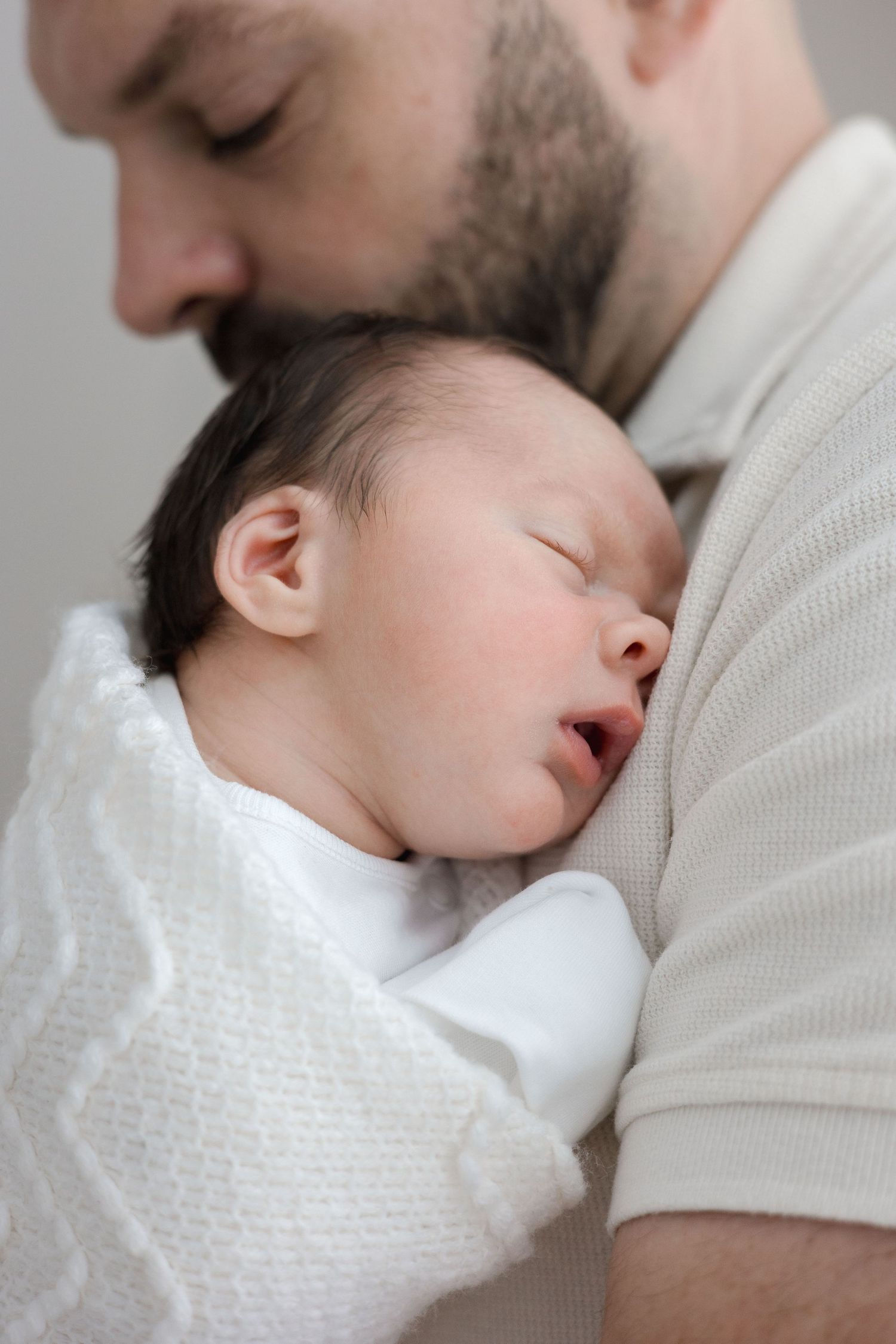 Bearded father tenderly kissing sleeping newborn baby wrapped in white knit blanket.