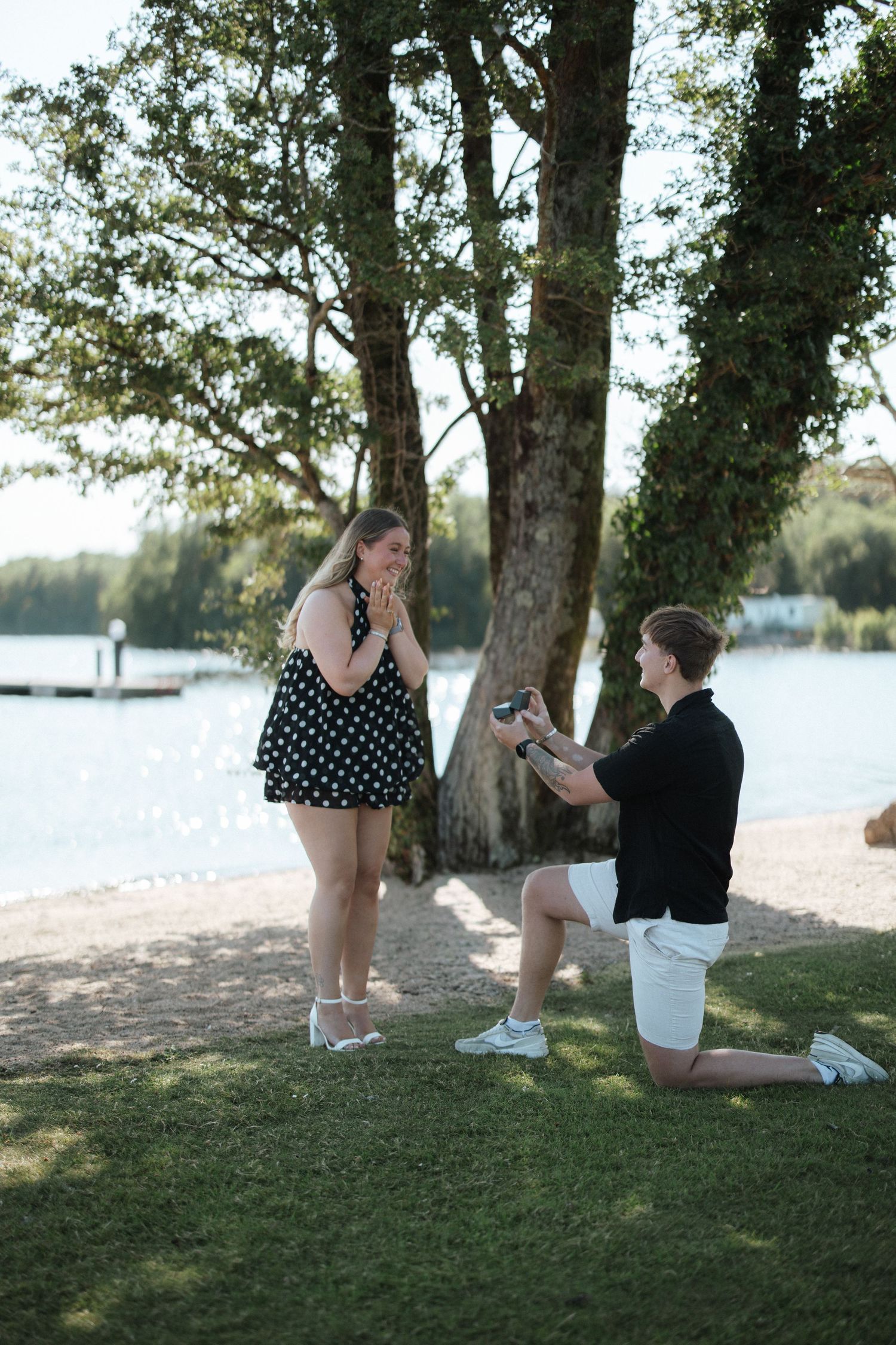 Man proposing on one knee to woman in polka dot dress by a lakeside, her hands clasped in surprise.