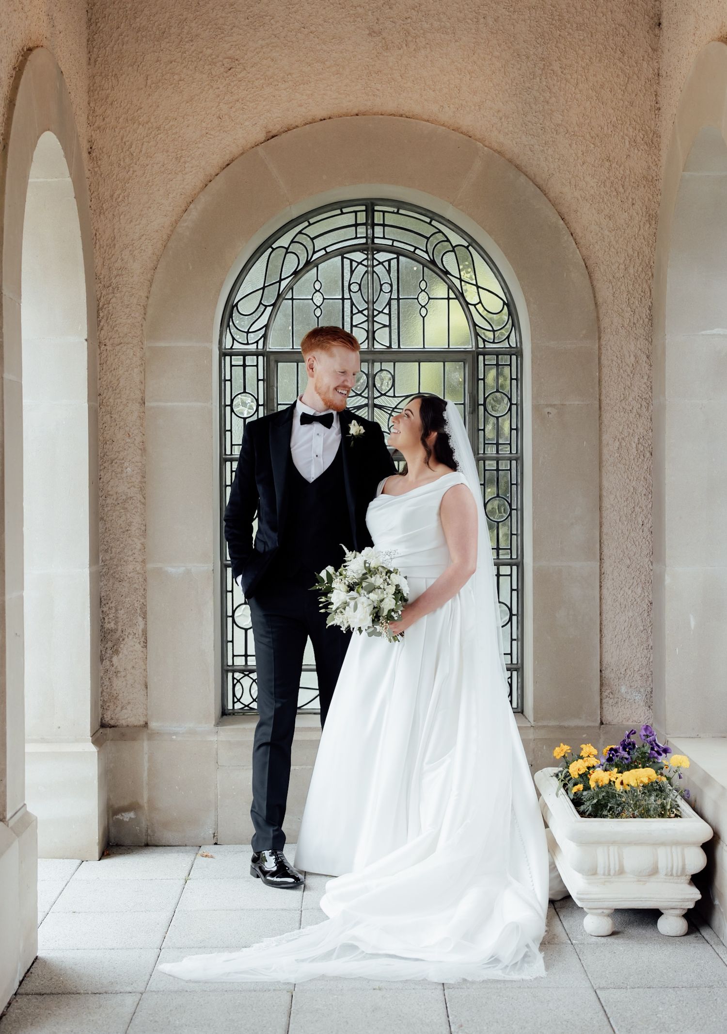 Bride and groom share a romantic moment under an elegant arched window with stained glass at their wedding venue.