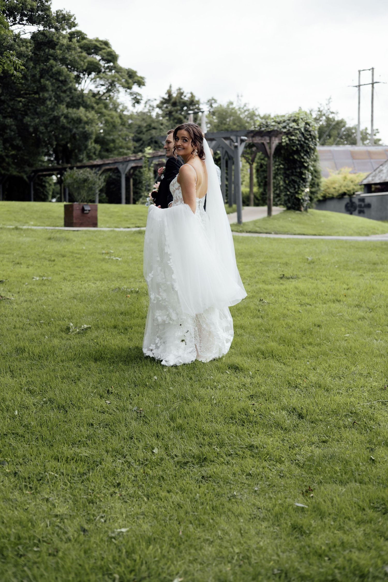 Bride in elegant white lace gown with long train standing on green lawn at wedding venue.