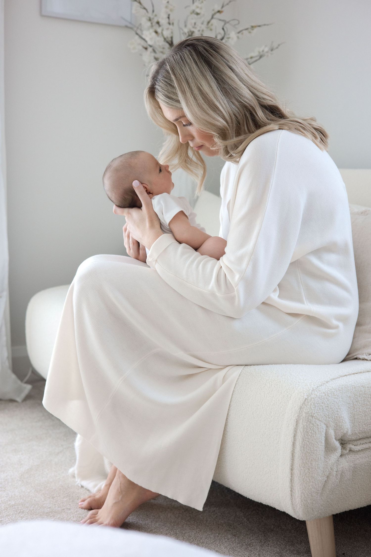 A blonde mother in a white dress sits on a bed, lovingly cradling her newborn baby.
