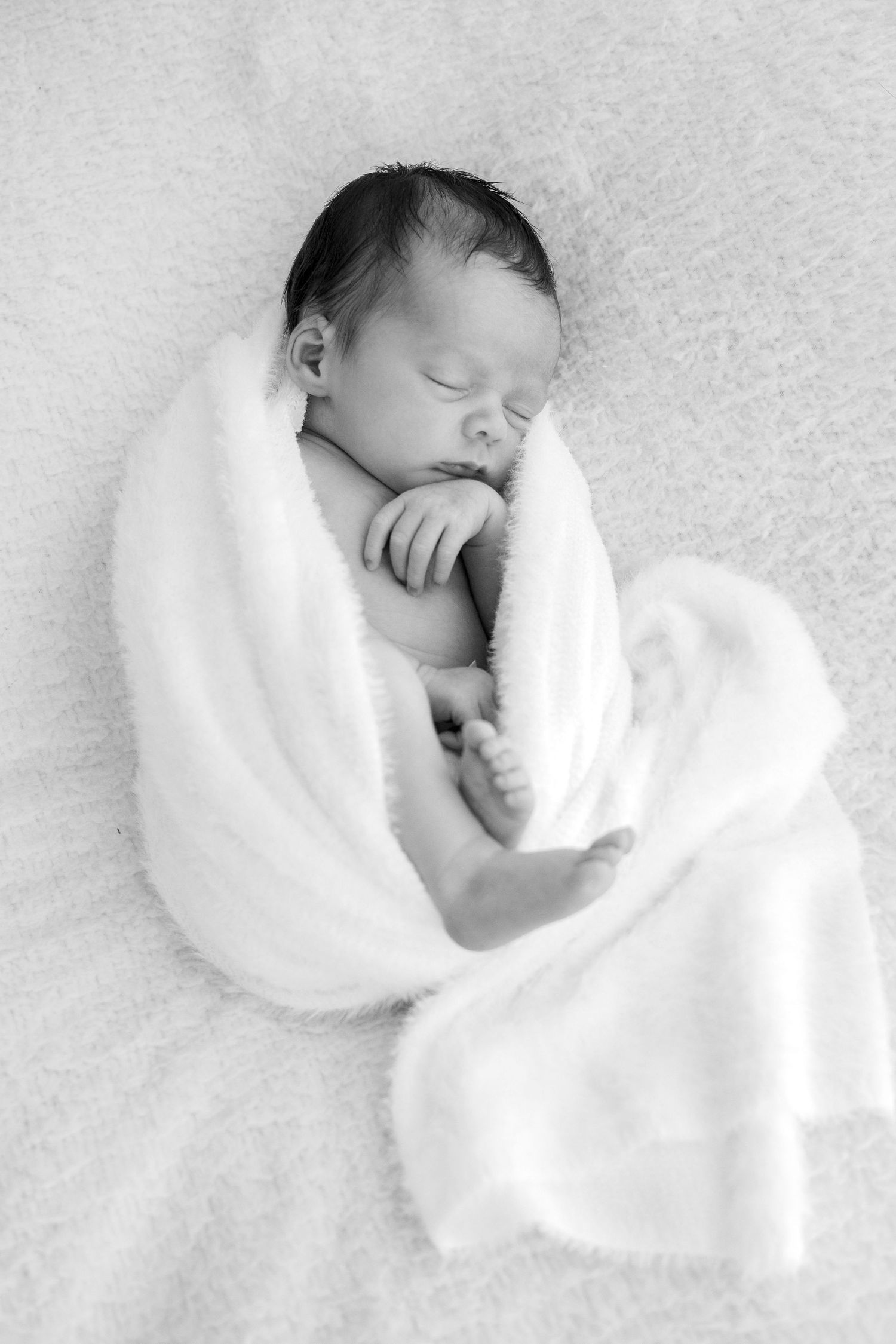 A peaceful newborn baby sleeps curled up in a white fluffy blanket in black and white.