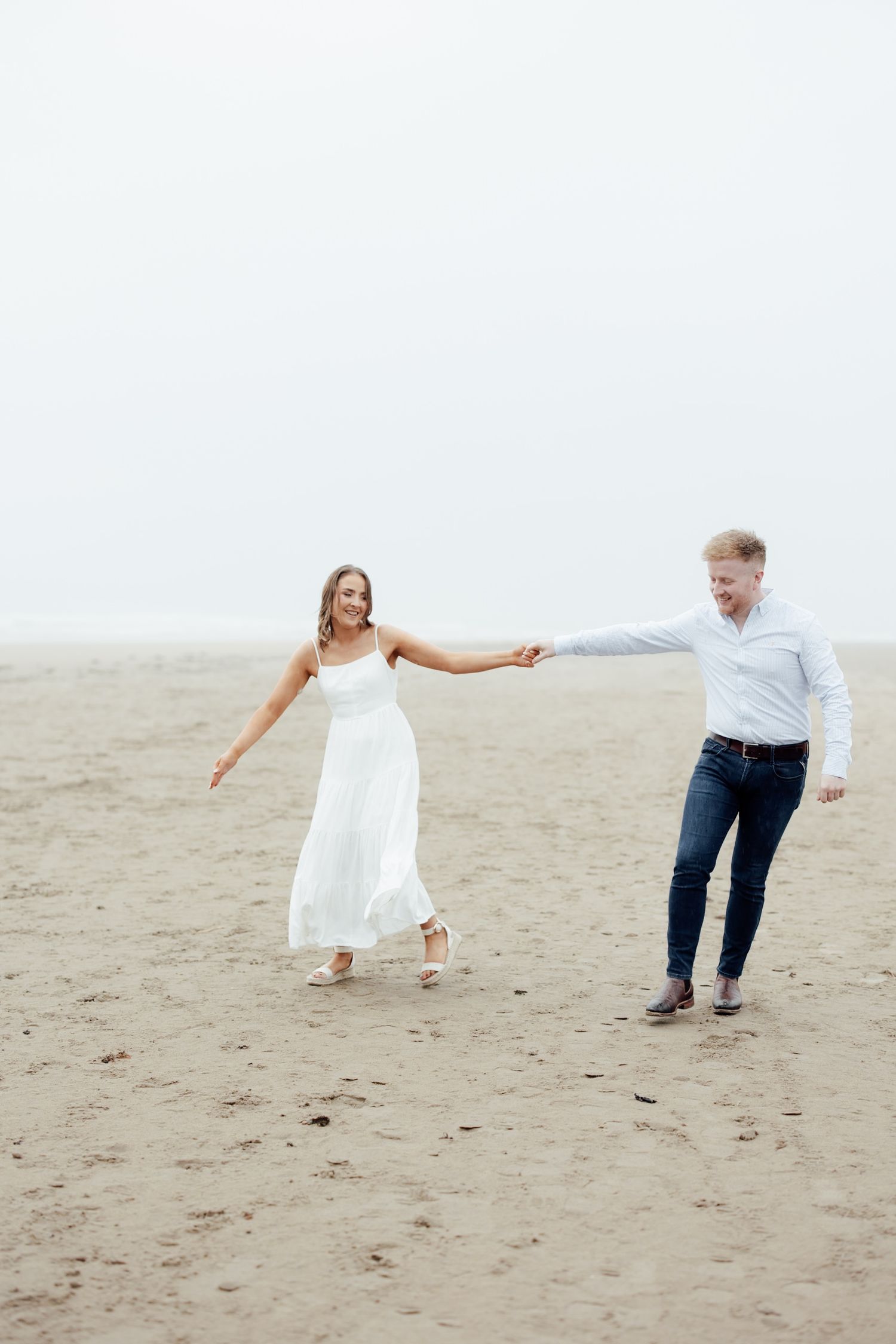 Couple holding hands and dancing on a sandy beach, woman in white dress, man in jeans and blue shirt.