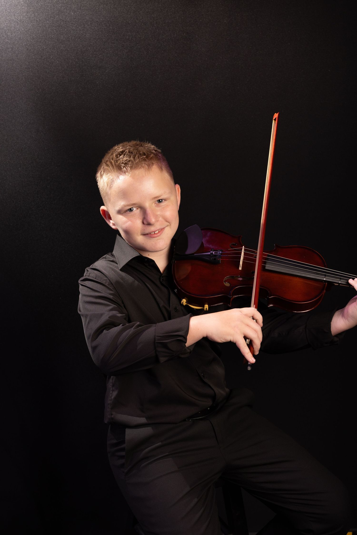 A violinist in formal black attire performs against a dark background while holding their instrument and bow.