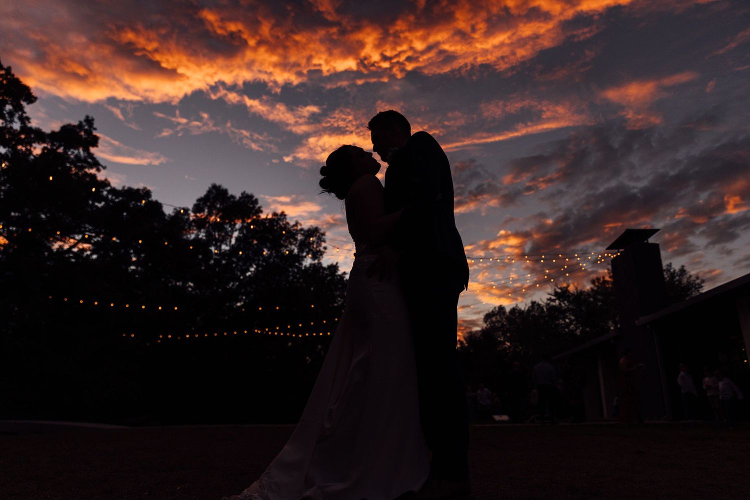 Silhouette of a bride and groom embracing against a stunning fiery sunset sky with string lights and trees.