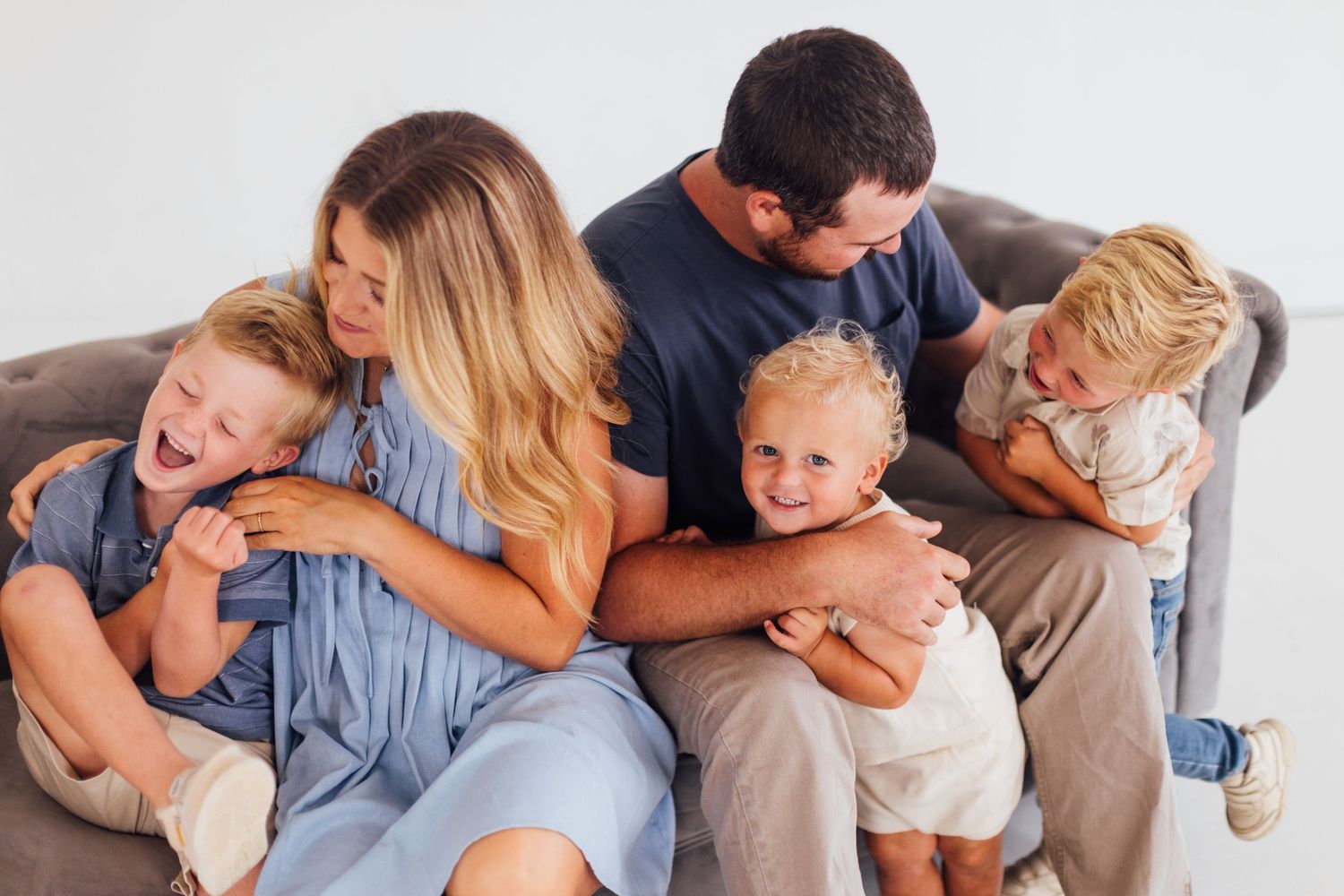 Happy family with three young blonde children laughing and cuddling together on a gray sofa during a photo session.