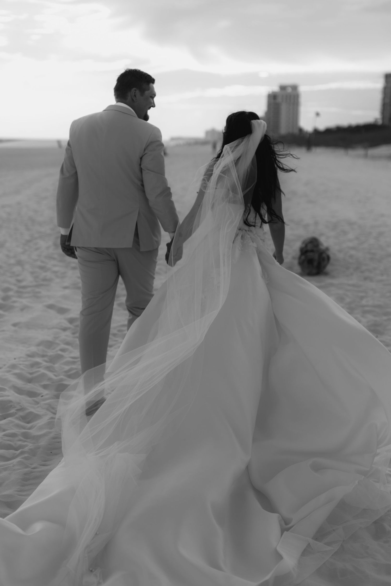 Black and white photo of a bride and groom walking hand in hand on a beach, her gown and veil flowing in the wind.
