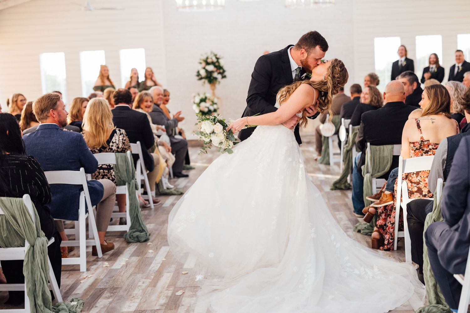 Bride and groom share a romantic kiss in the aisle during their elegant indoor wedding ceremony with seated guests.